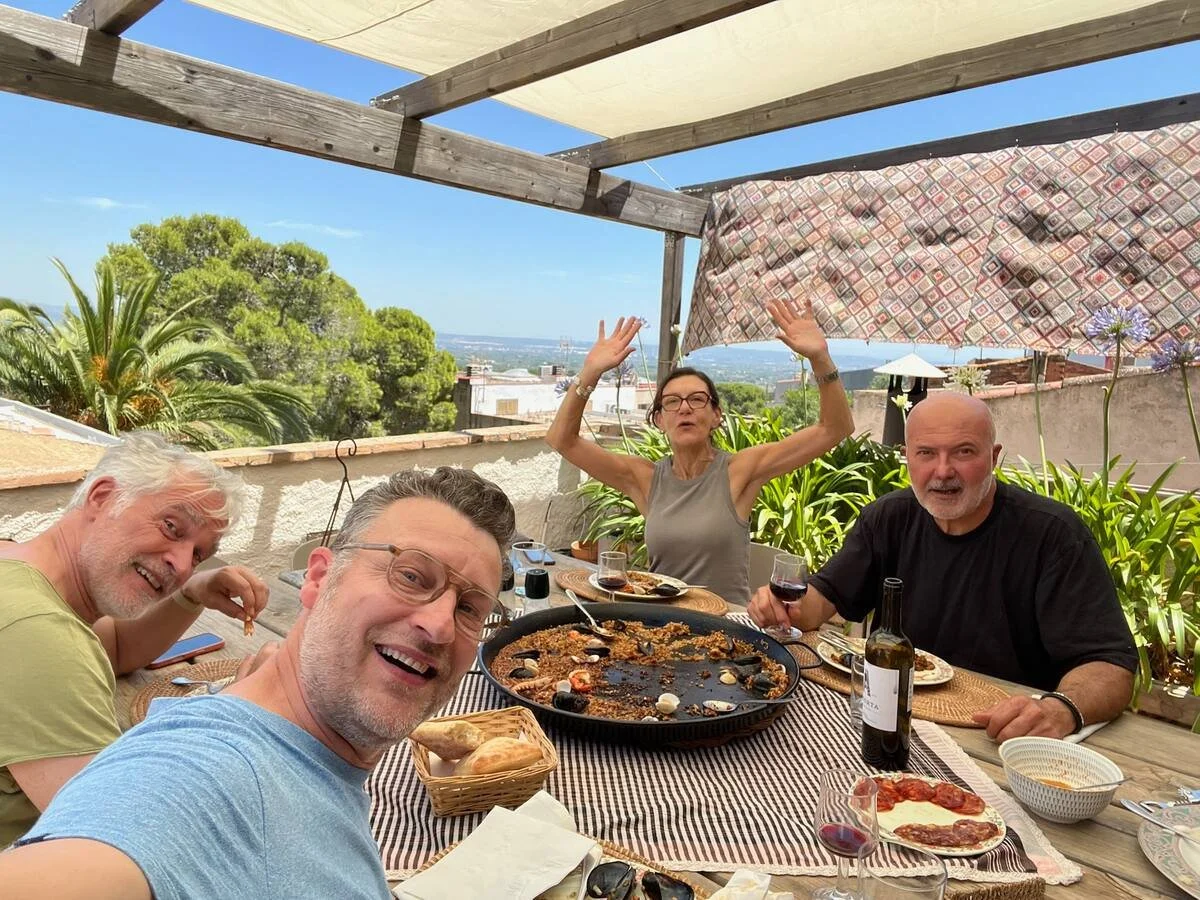 Cuatro personas en una terraza celebrando con comida y vino tinto, rodeadas de plantas y con vista al cielo y la ciudad.