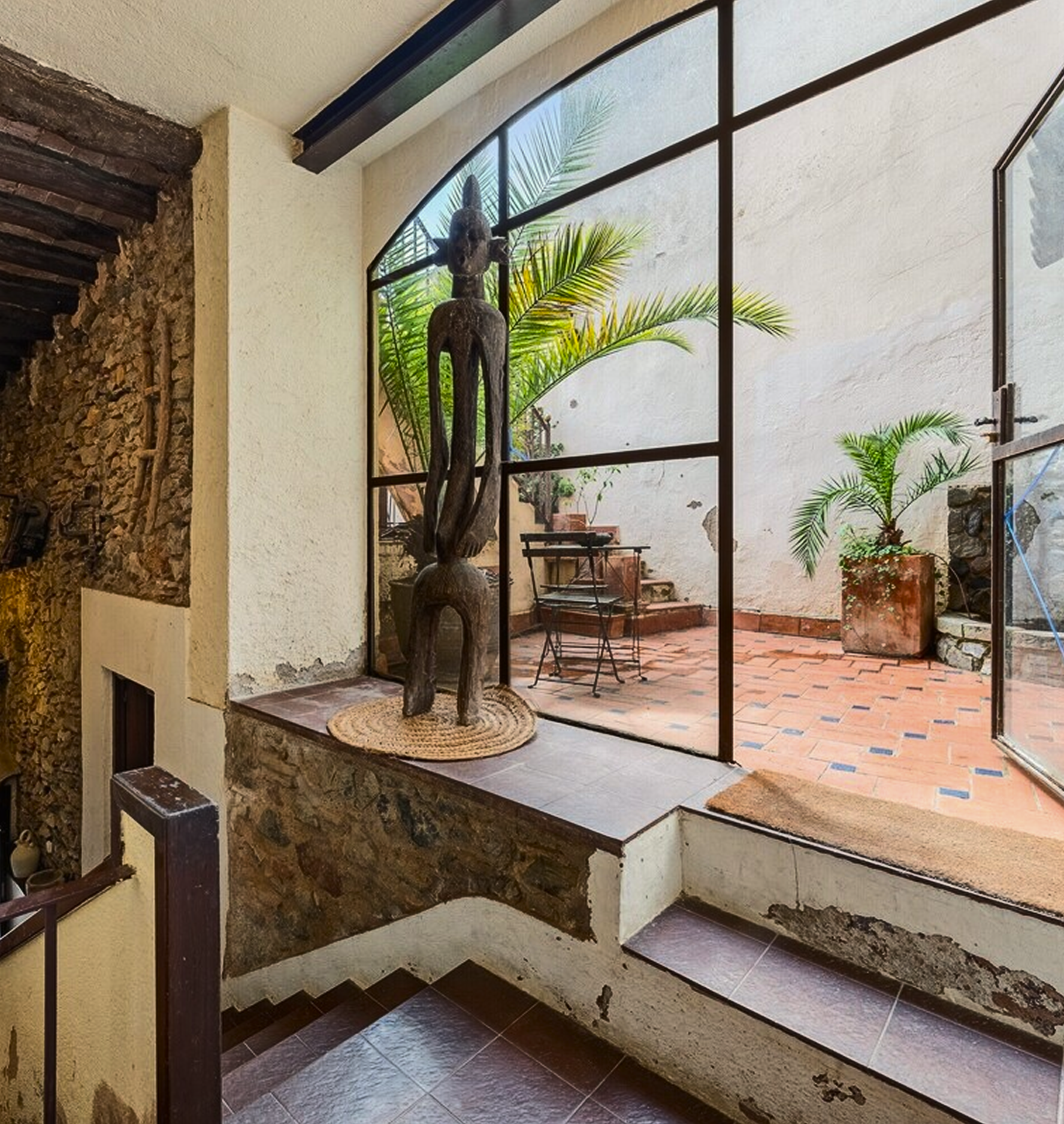 Interior de una casa con una escultura de madera africana en una repisa, una pared de piedra, plantas en macetas y una puerta de vidrio que da al patio con baldosas de terracota.