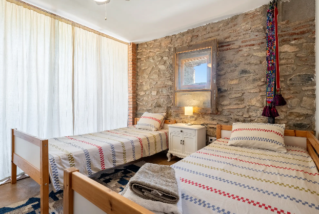 Habitación con dos camas individuales, una pared de piedra, cortinas blancas, y una lámpara sobre una mesita de noche entre las camas.