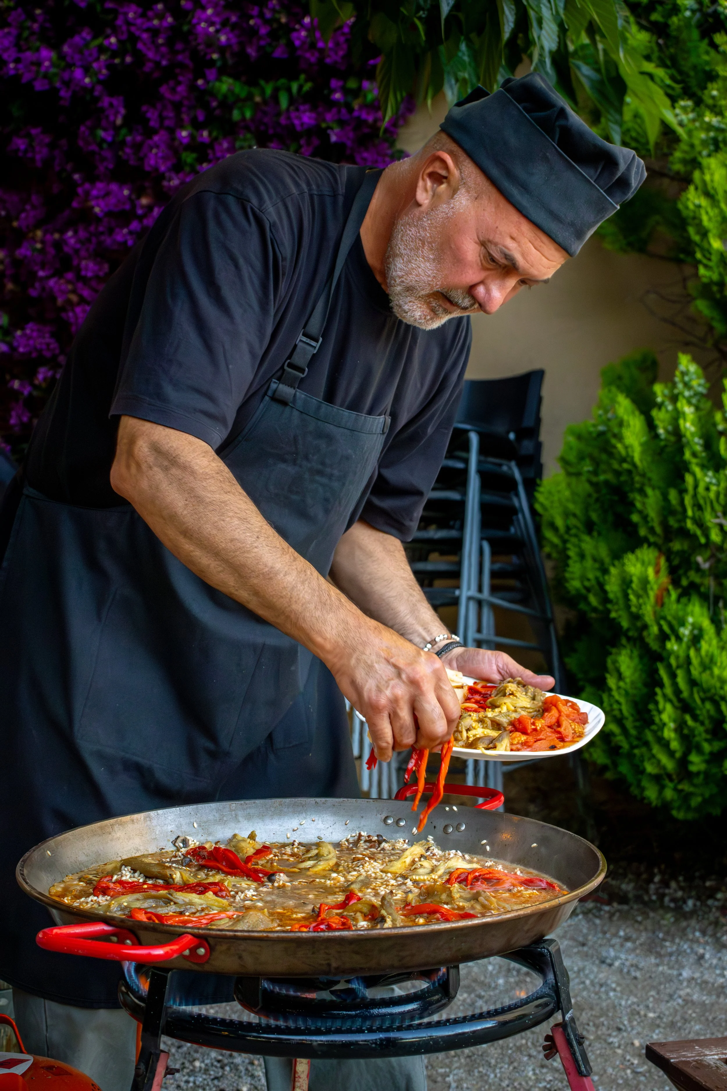 Un chef prepara y sirve comida en una plancha grande, con vegetales y carne, en un entorno al aire libre con plantas y flores de fondo.