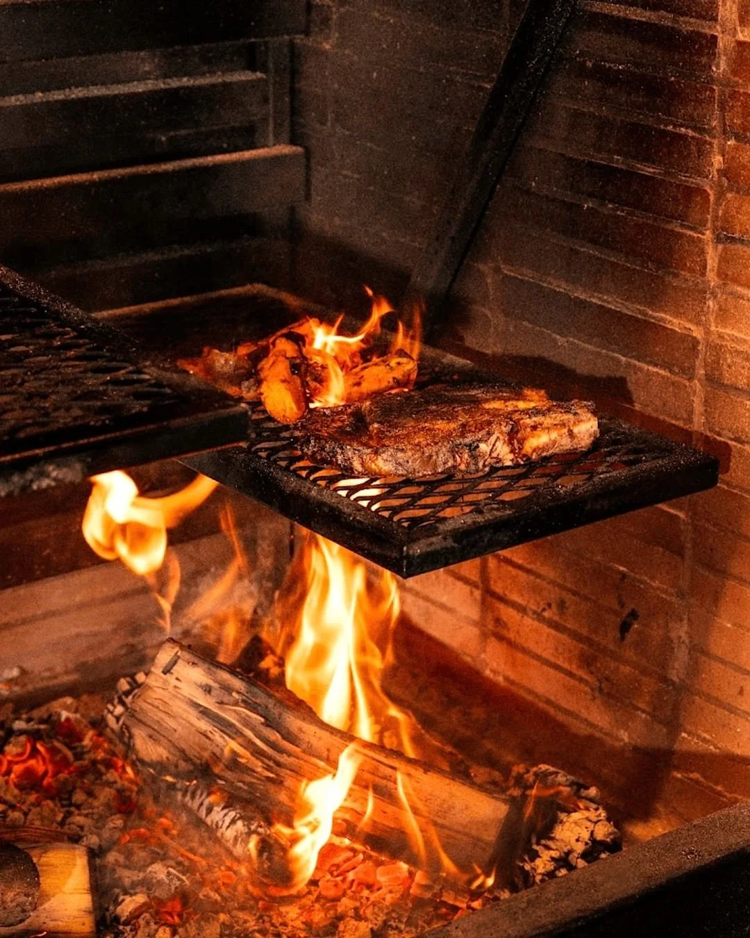 Carne asada en una parrilla de carbón con llamas ardientes y madera en las brasas.