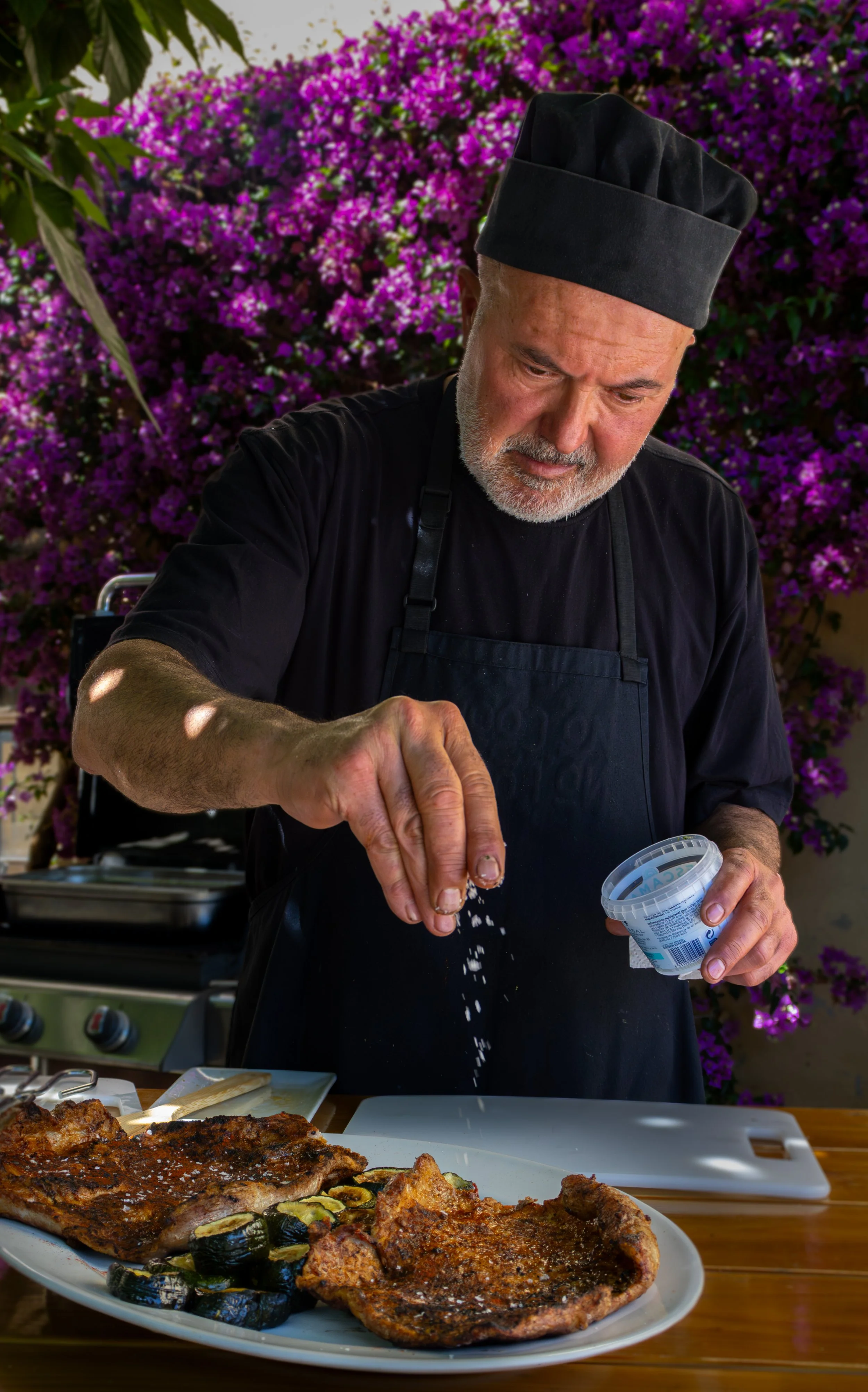 Hombre cocinero rociando sal en una parrilla con comida, en fondo de flores moradas.