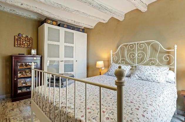 Cama de matrimonio con cabecera de hierro blanco, ropa de cama con estampado floral, una lámpara de mesa encendida, armario blanco con puertas de vidrio, y un mueble de madera oscura con objetos decorativos en una habitación con paredes beige y techo con vigas de madera blancas.