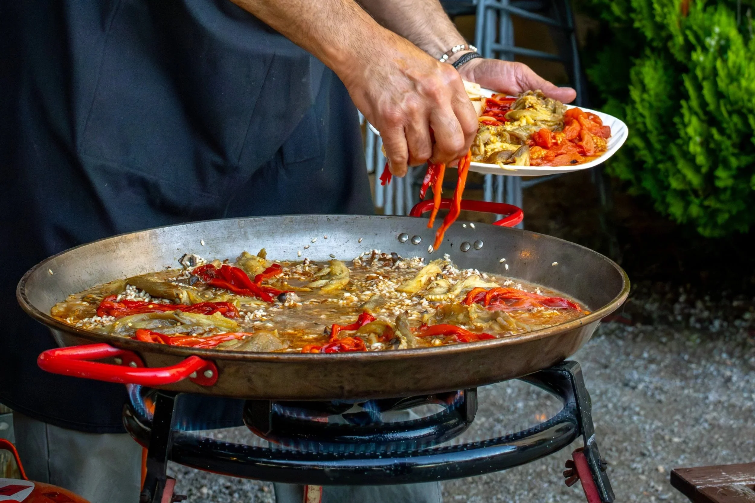 Persona cocinando un plato en una sartén grande, usando una espátula para mezclar ingredientes como pimientos rojos y vegetales en una cocinera exterior con fondo de plantas verdes.