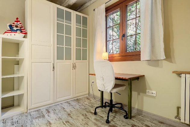 Habitación con escritorio, silla, armario y estantería junto a una ventana con cortinas blancas.