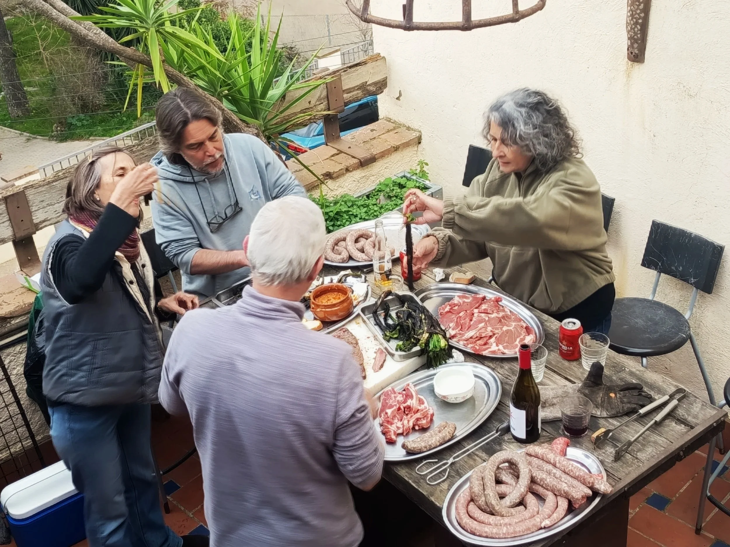 Grupo de personas sirviéndose carne y acompañamientos en una comida al aire libre, con vegetación y paredes de fondo.