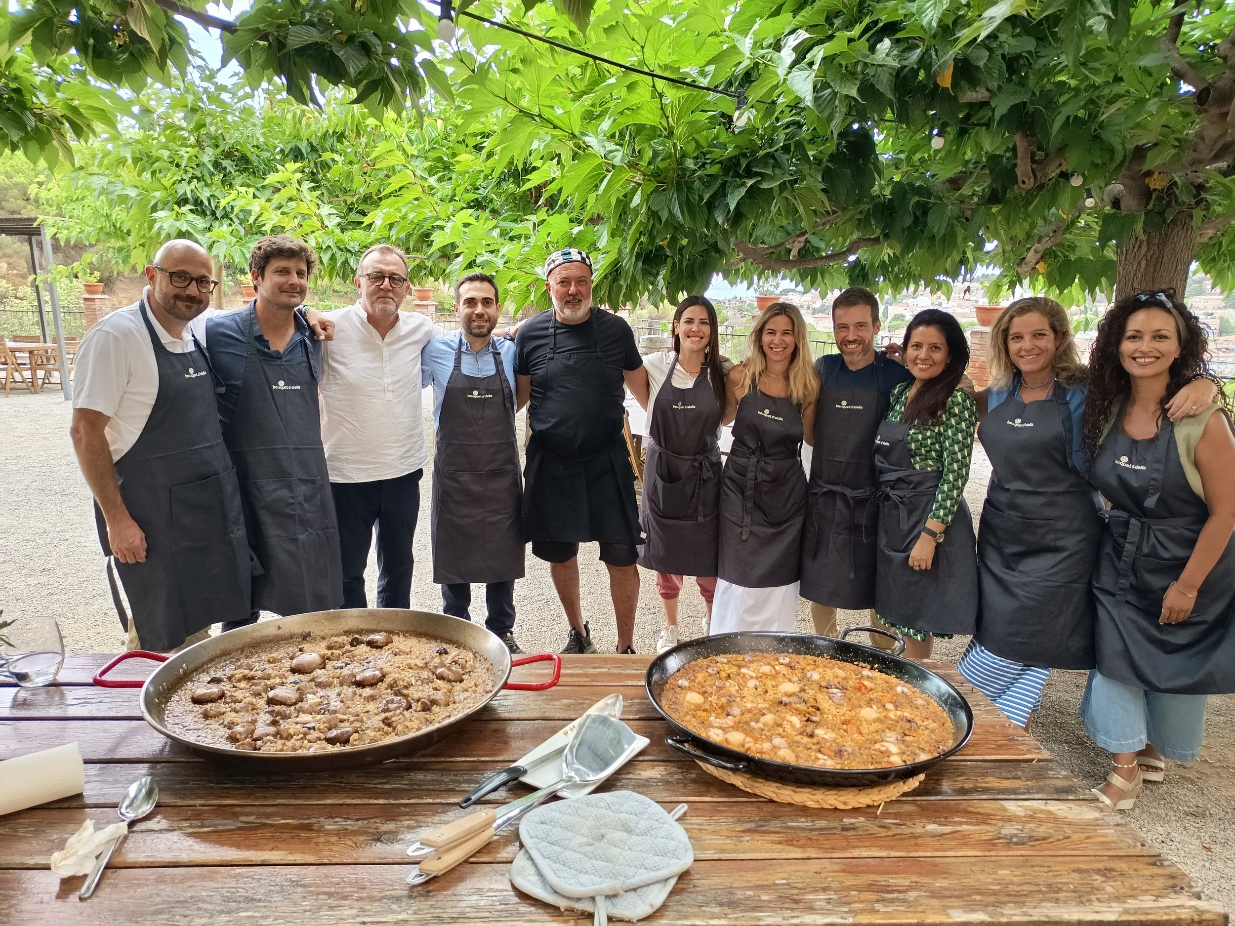 Grupo de personas sonriendo y posando en un entorno al aire libre debajo de un árbol, con platos de comida en una mesa de madera en primer plano.