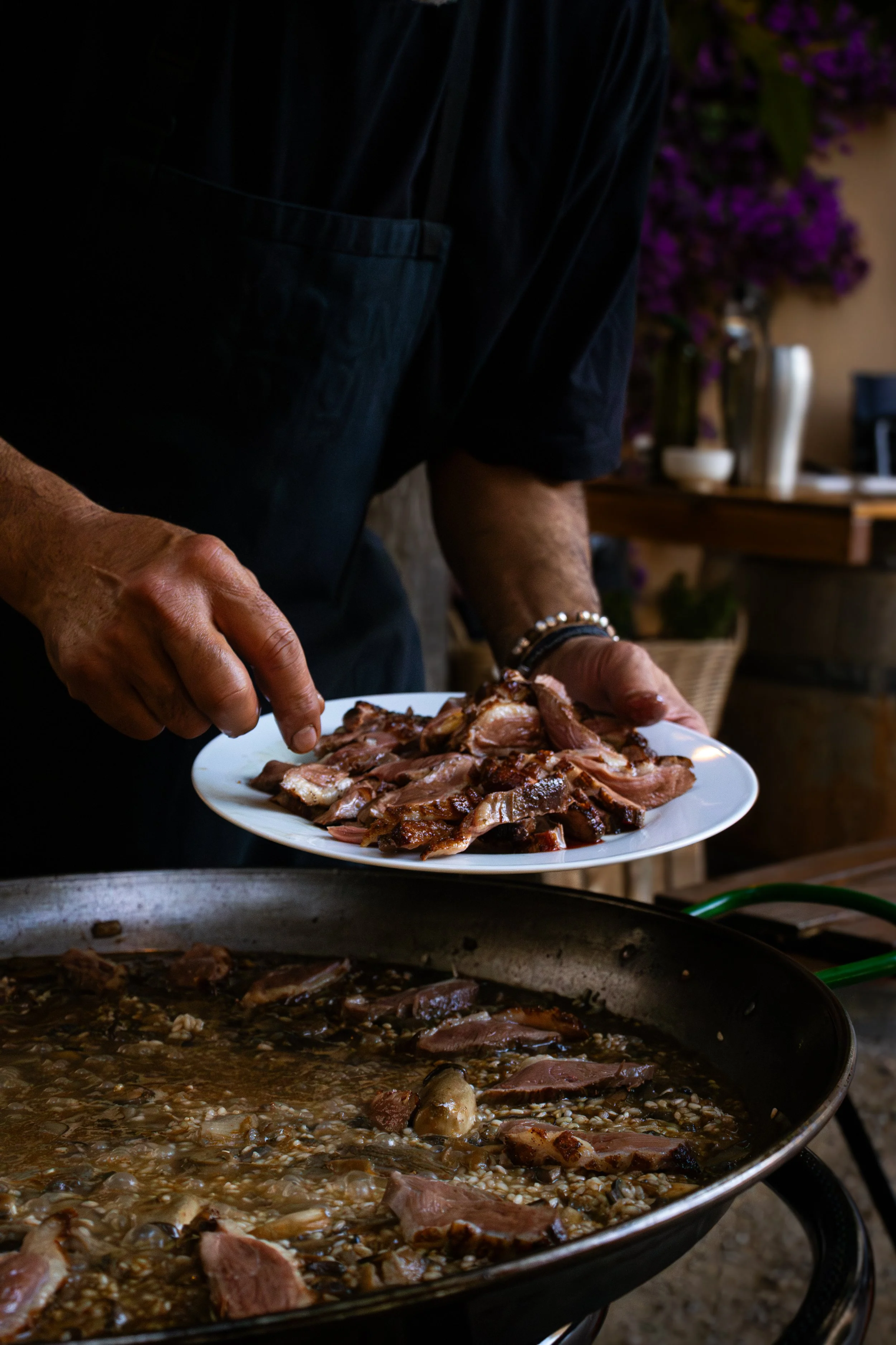 Persona sirviendo carne en un plato, con una olla con arroz y carne cocida en primer plano y flores moradas al fondo.