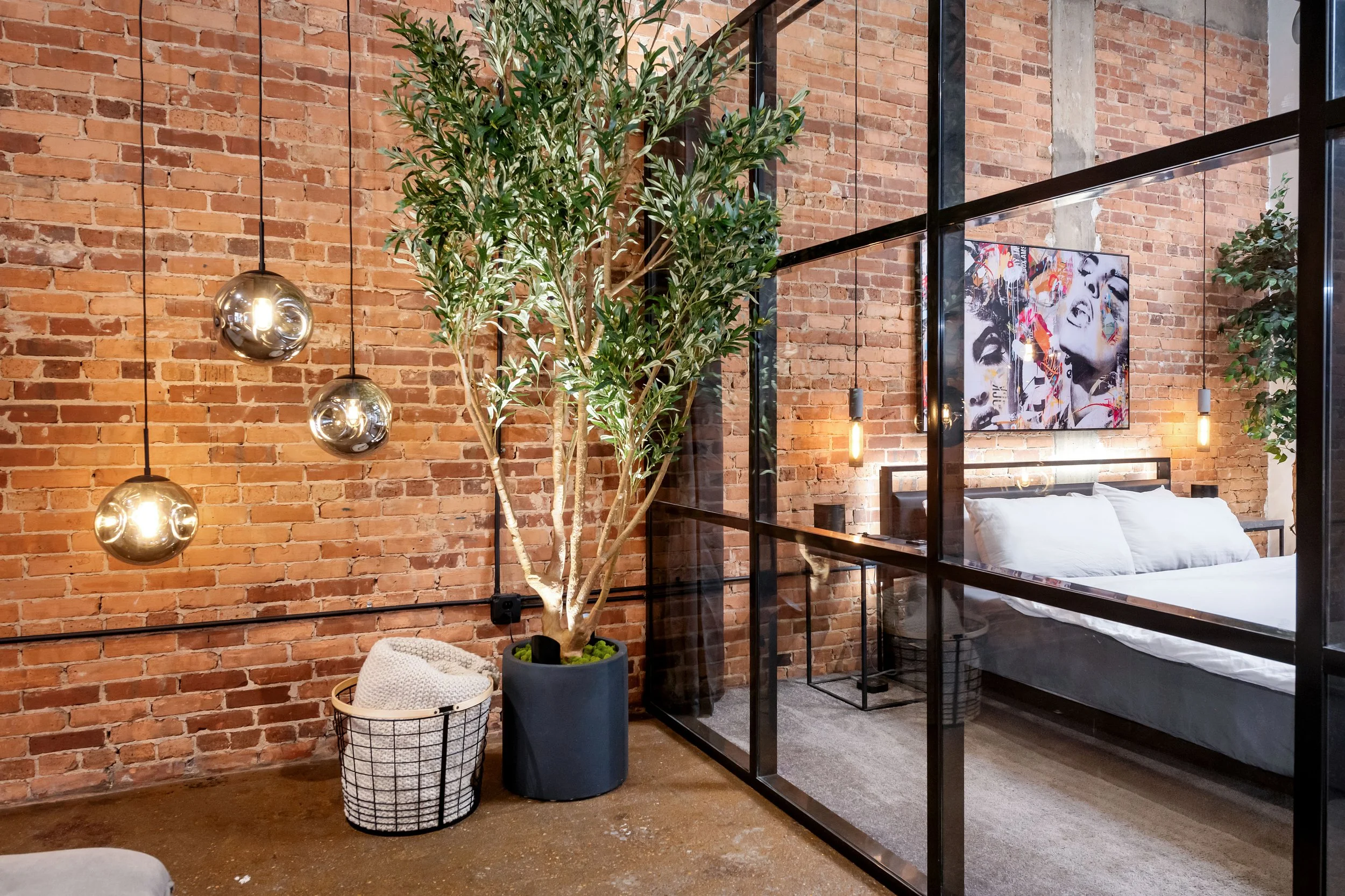 Interior of a modern bedroom with exposed brick wall, a potted plant, stylish hanging lights, and a framed black and white pop art poster on the wall.