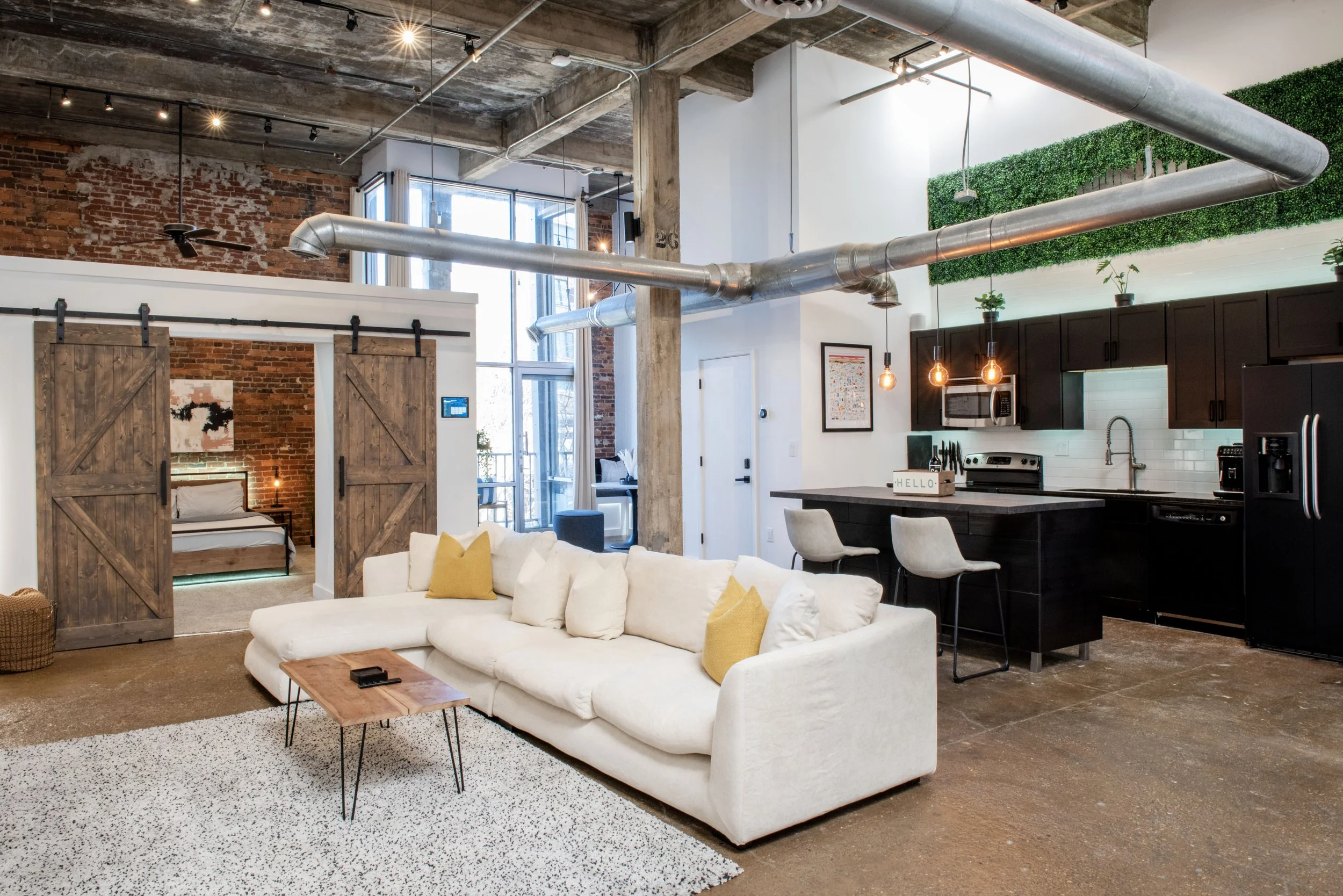 Open-concept loft apartment with exposed brick walls, large windows, modern kitchen with black cabinets, white subway tile backsplash, and bar seating. Living room features a white sectional sofa with yellow pillows, a wooden coffee table, and a text