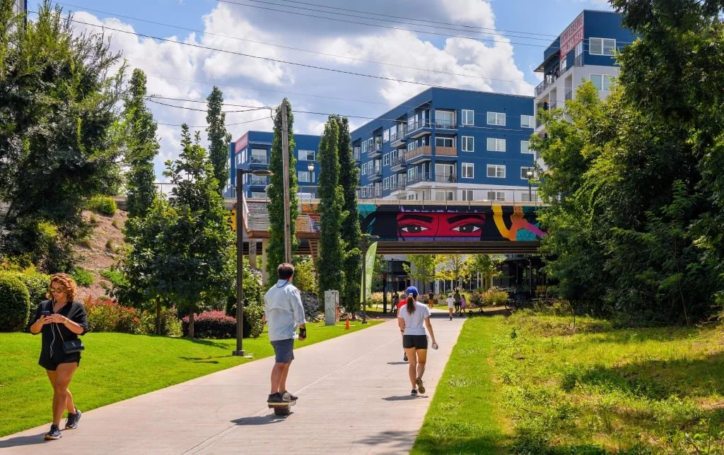 People walking and skateboarding on a park pathway with trees and grass, modern building with colorful mural in the background.