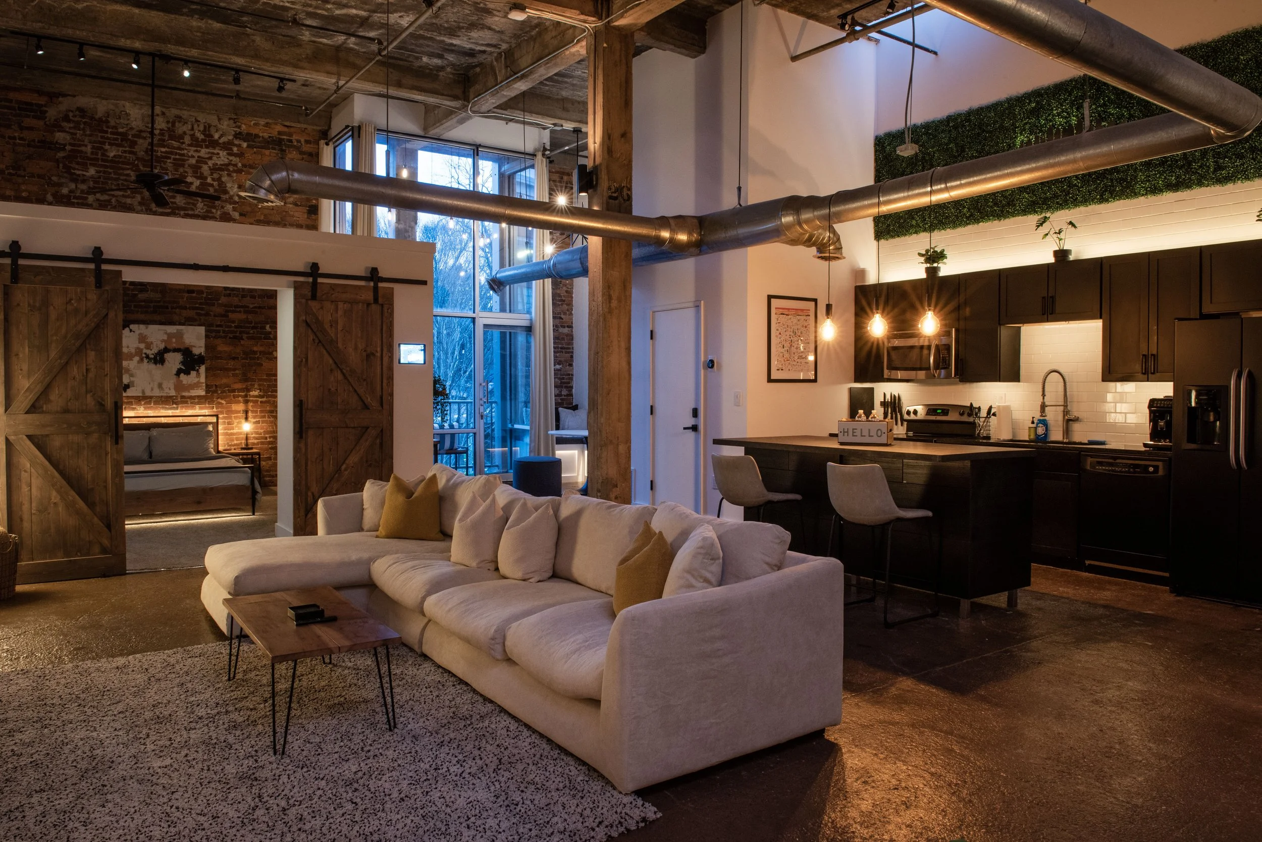 Modern loft apartment interior with exposed brick walls, large windows, a cozy living room with a white sectional sofa and yellow pillows, and a visible kitchen area with black cabinets, a countertop with a sign that says 'HELLO', and hanging pendant