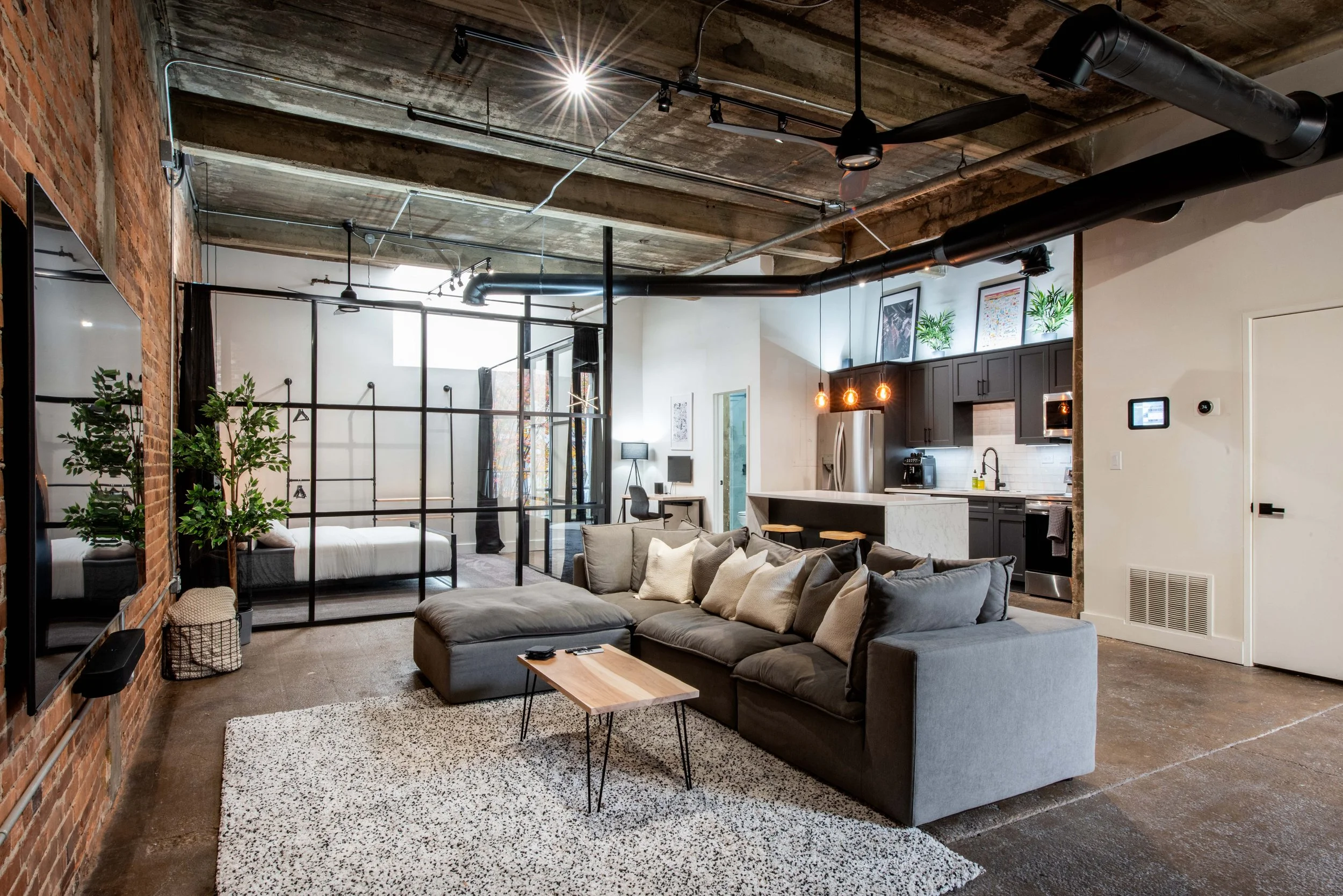 A modern, industrial-style apartment living room with a gray sectional sofa, a beige armchair, a small wooden coffee table, and a patterned rug. The room features exposed brick walls, a concrete ceiling with visible ductwork, large windows with black curtains, and a kitchen area in the background with black cabinets, a stainless steel refrigerator, and pendant lights.