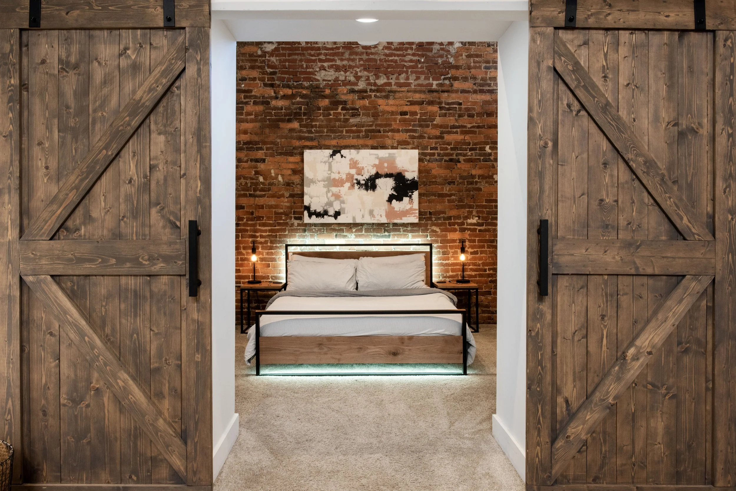 A bedroom with an exposed brick wall, a bed with white bedding, and bedside tables with lamps, viewed through sliding barn doors made of wood.