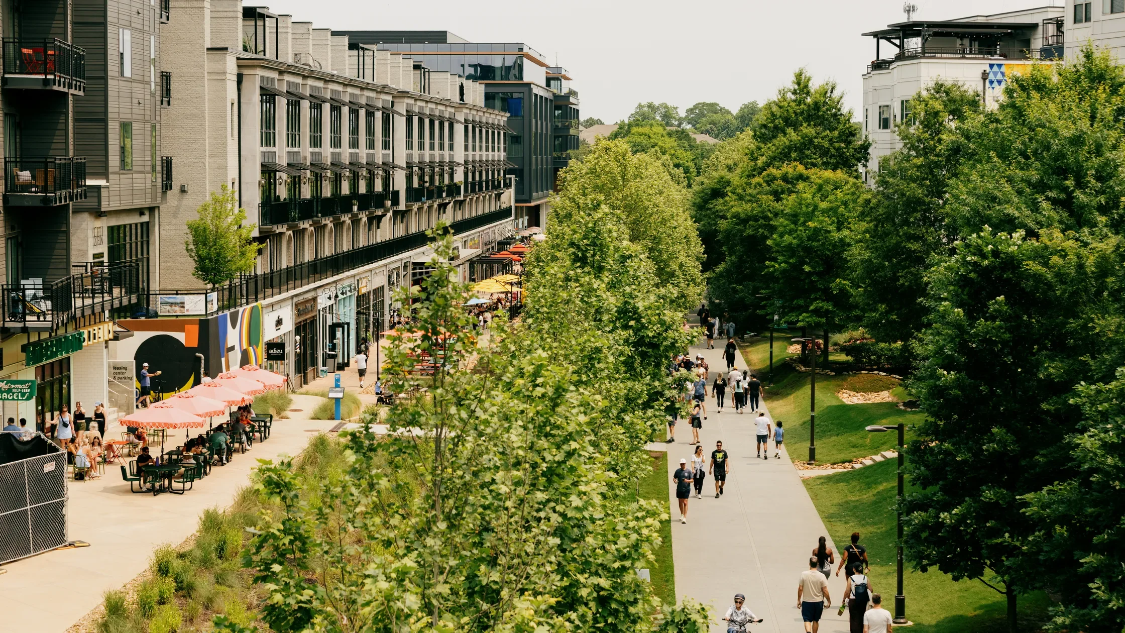 A lively city street with multi-story buildings, outdoor cafes with orange umbrellas, and people walking on the sidewalk lined with trees and green lawns.