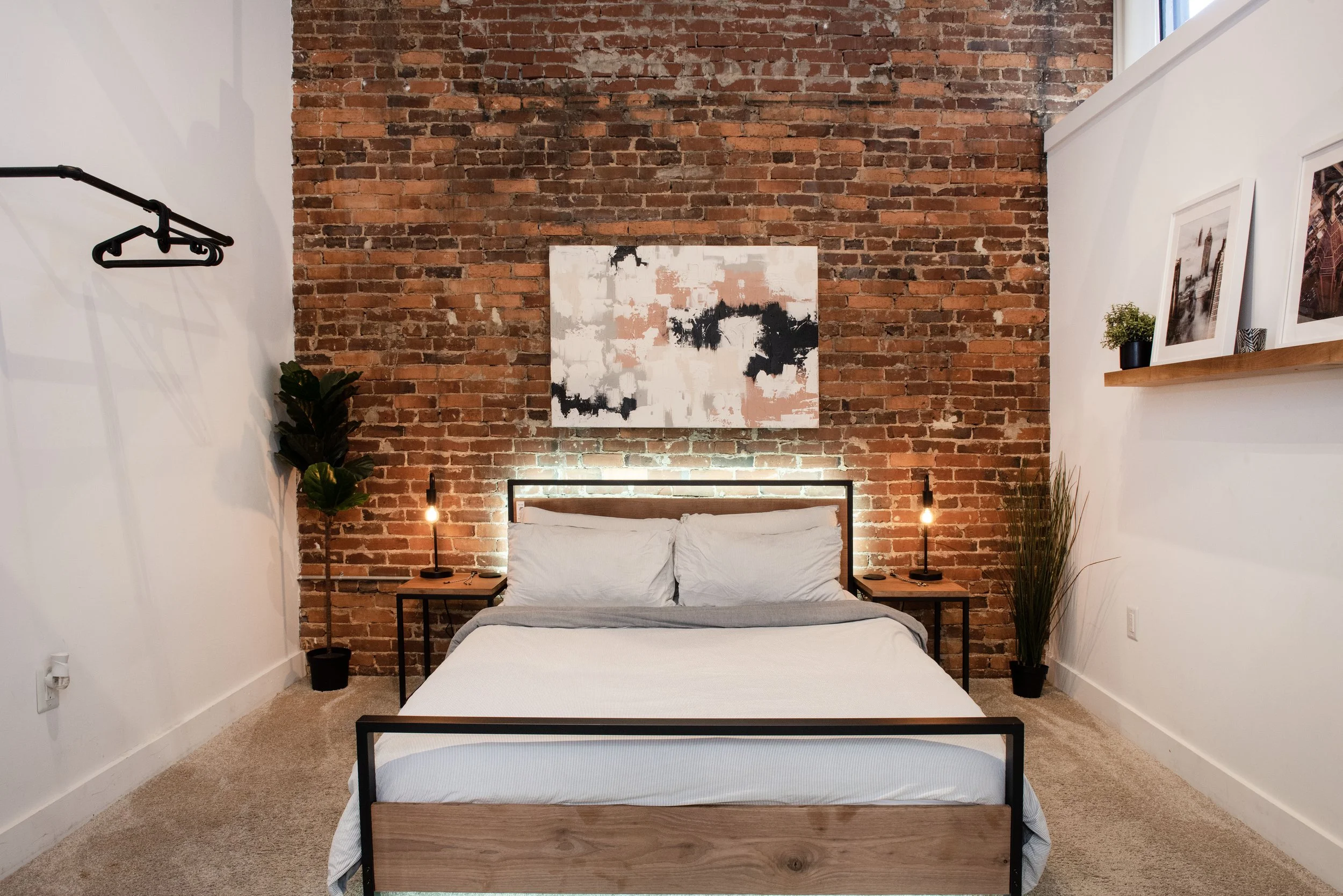 A bedroom with a brick accent wall behind the bed, featuring a modern abstract painting. There are two small nightstands with lamps, a potted plant on the right shelf, and a clothing rack on the left wall. The bed has white bedding and pillows, with 