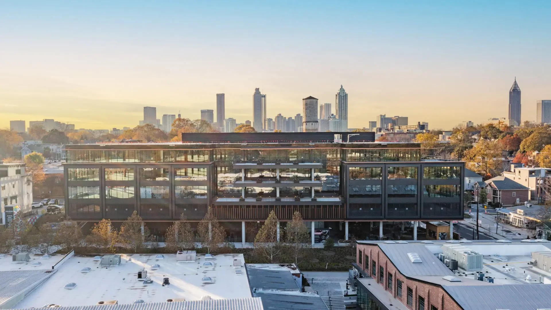 City skyline at sunset with tall buildings in the background, modern glass building in the foreground, surrounded by trees and smaller structures.