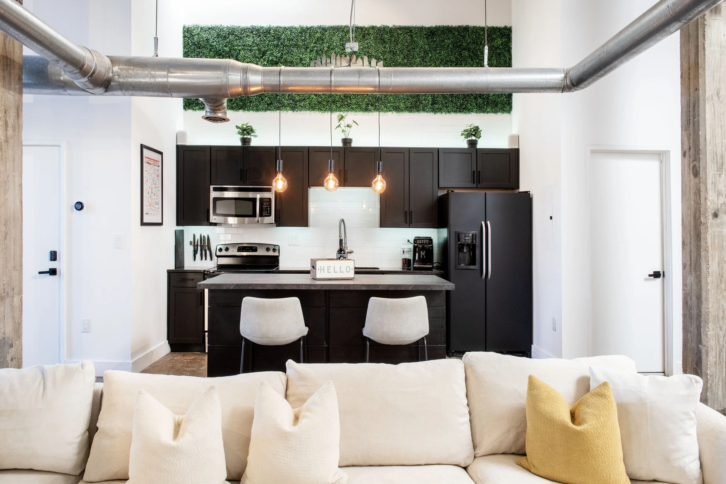 View of a modern kitchen with black cabinets, stainless steel appliances, and hanging Edison bulb lights above a kitchen island. There is a green artificial plant wall on the upper part of the back wall and potted plants on top of the cabinets. Part 