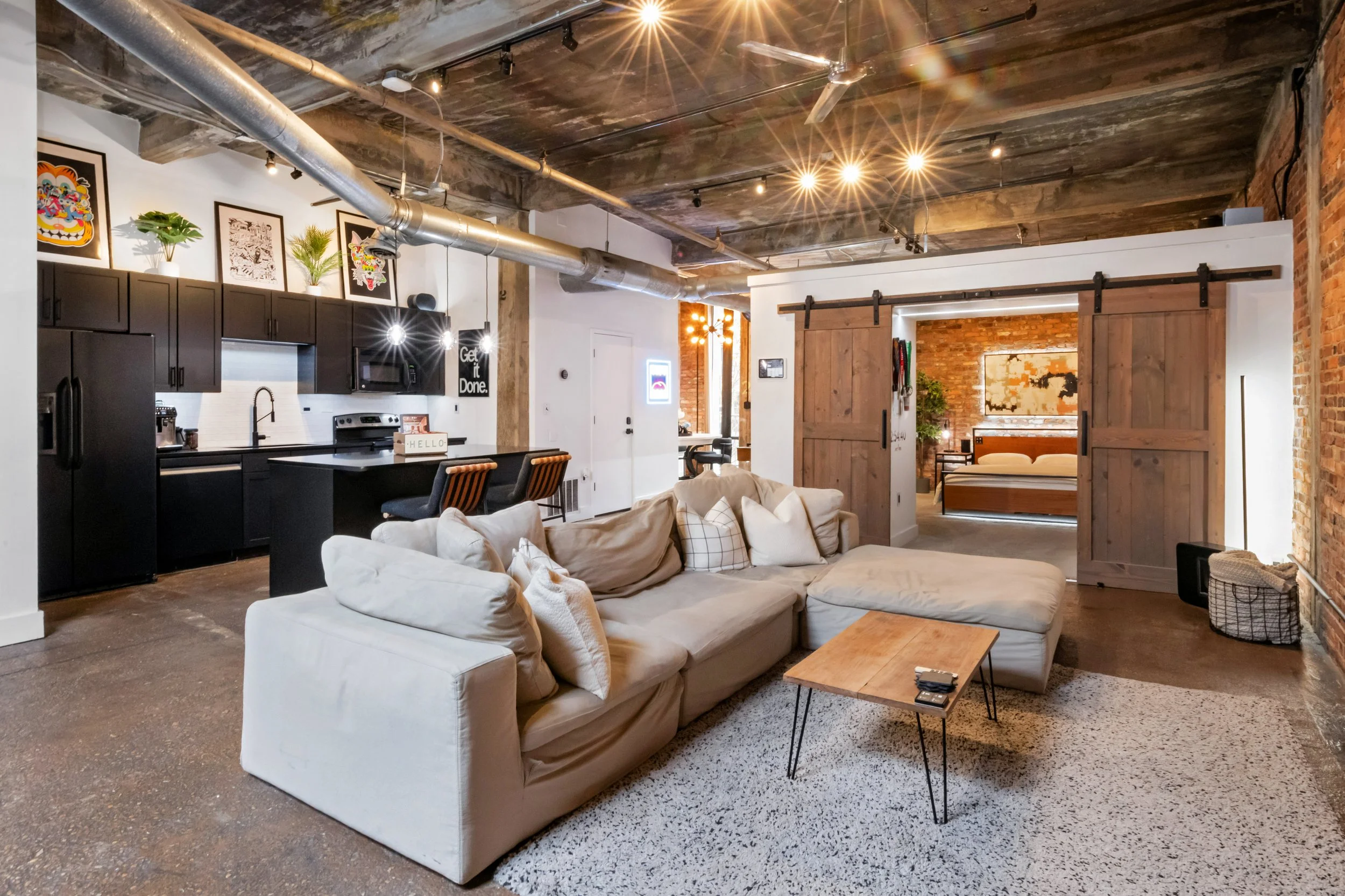 Open living space with exposed brick walls and wooden sliding barn doors leading to a bedroom. The space features a white sectional sofa with multiple cushions, a wooden coffee table, and a cozy area rug. The kitchen area has dark cabinetry, a white backsplash, and modern appliances, with framed artwork and potted plants on top. The ceiling has an industrial look with exposed ductwork and track lighting, along with a ceiling fan.