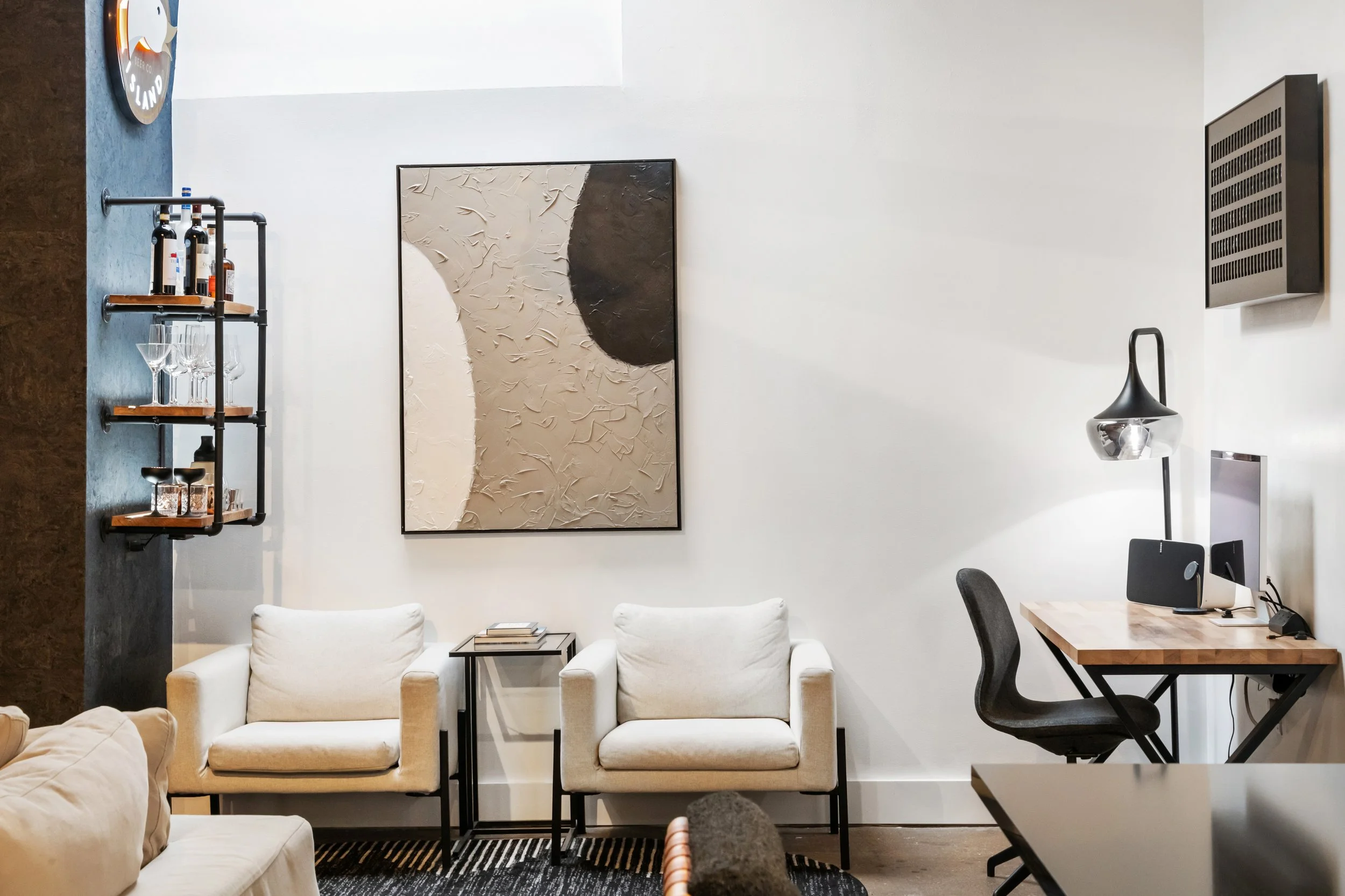 Modern living room with white sofas, a black framed abstract art piece on a white wall, a bar shelf with bottles and glasses on the left, a small side table with books in front of the sofas, a black desk lamp on a wooden table on the right, and a com