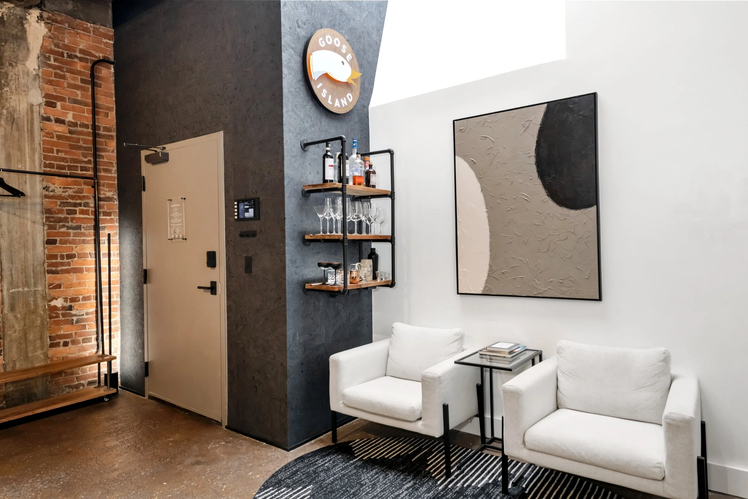 Modern interior space with exposed brick wall, dark textured wall with a circular 'Goose Island' logo, wooden shelves with bottles and glassware, large abstract painting, white chairs, and a black table.