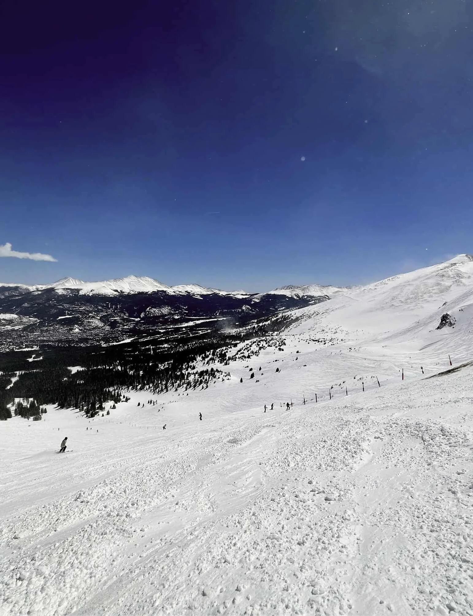 Snow-covered mountain landscape with skiers and snowboarders on a groomed slope, under a clear blue sky with distant mountain peaks.