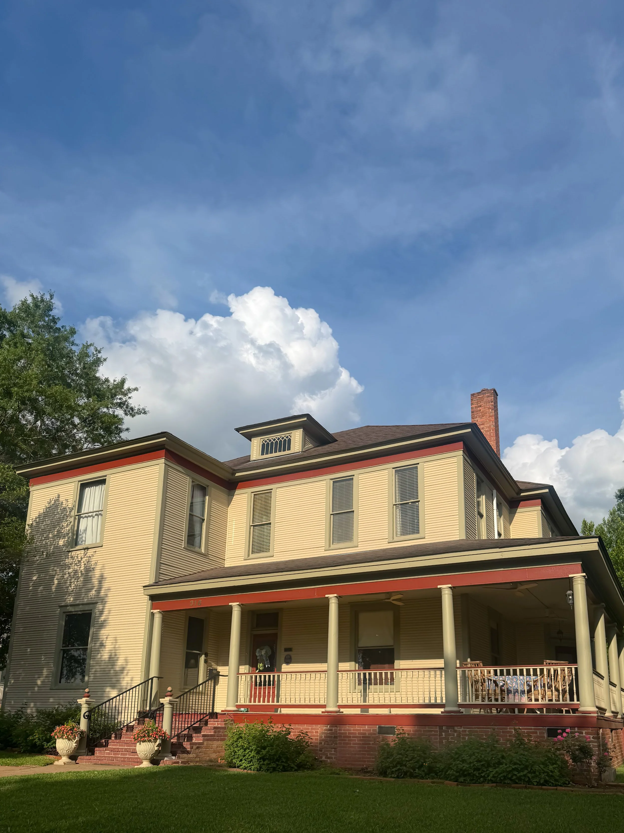 A large, historic, yellow house with red trim and a covered front porch, surrounded by a well-kept lawn and garden with potted flowers, under a partly cloudy blue sky.