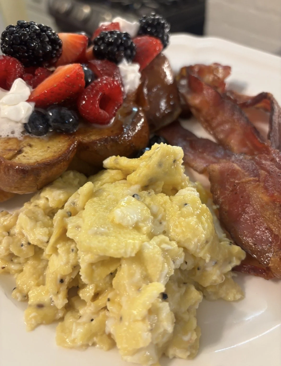 Breakfast plate with scrambled eggs, bacon, roasted potatoes topped with strawberries and blackberries, and dollops of whipped cream.