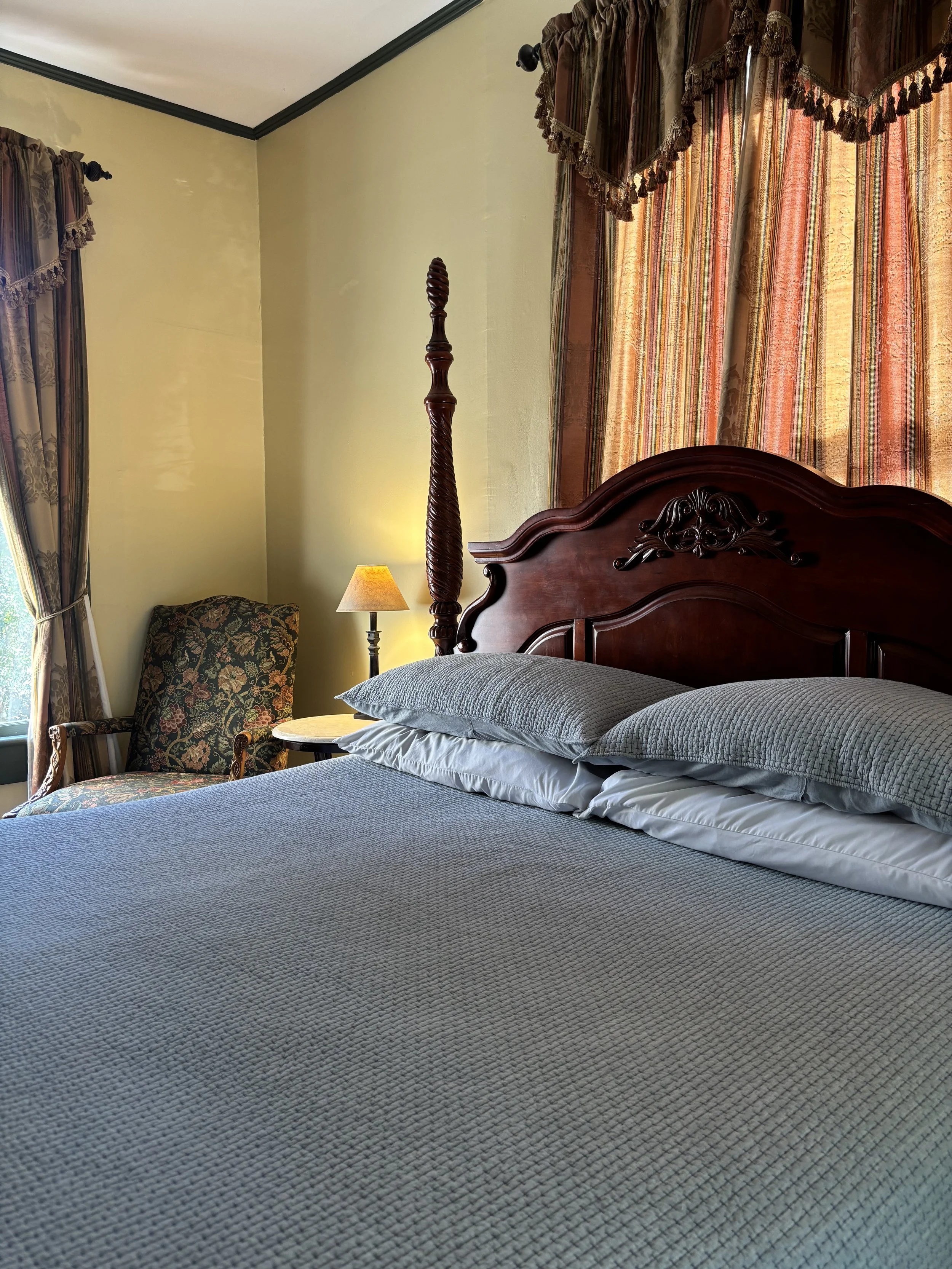 A bedroom with a neatly made bed, gray bedding, and a dark wooden headboard. To the left, there's a tall wooden bedpost, an armchair with floral upholstery, a small table, a table lamp, and curtains with stripes and tassel trim.