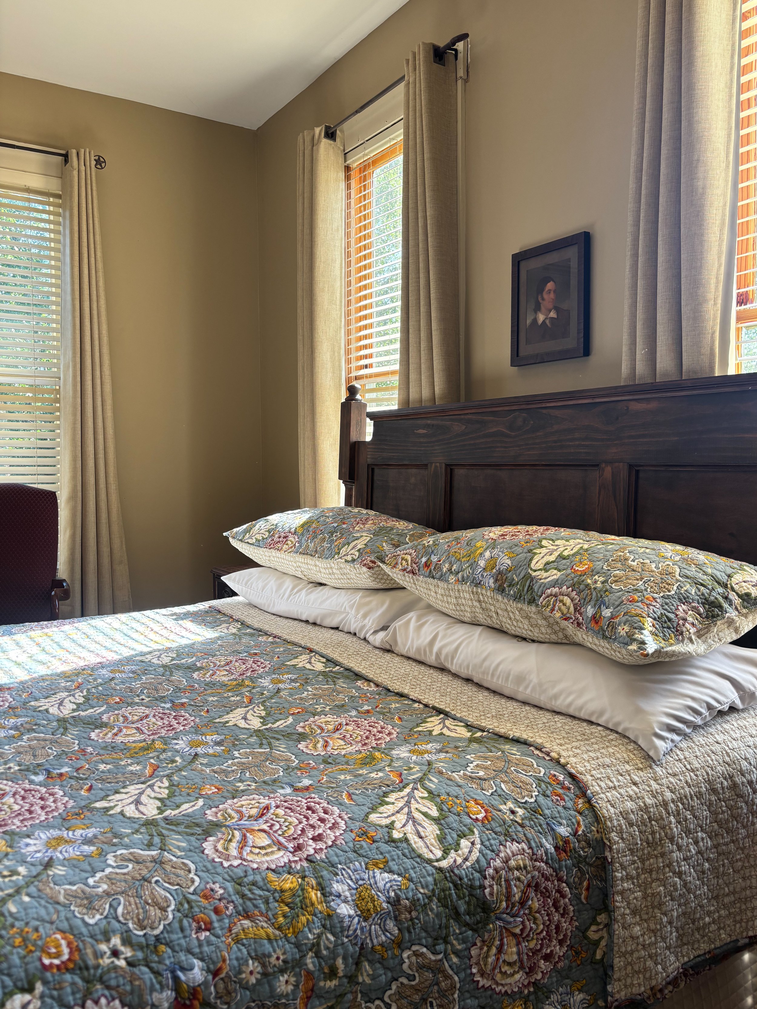 A bedroom with a wooden bed frame, patterned quilt, and two pillows, with sunlight coming through the windows which have beige curtains and wooden blinds, and a framed portrait on the wall.