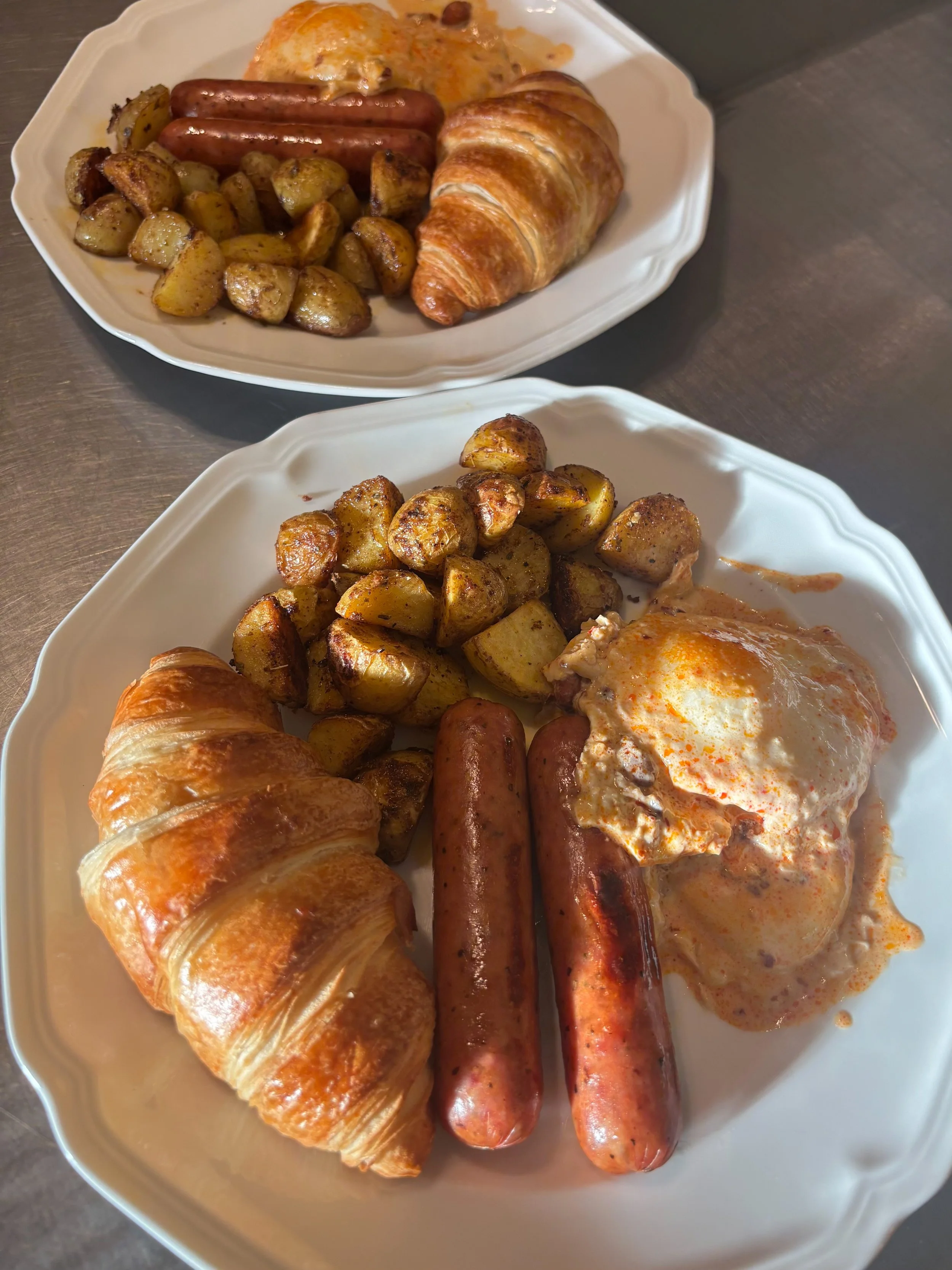 Plate with roasted potatoes, sausages, croissant, and fried eggs, served on white dishes.