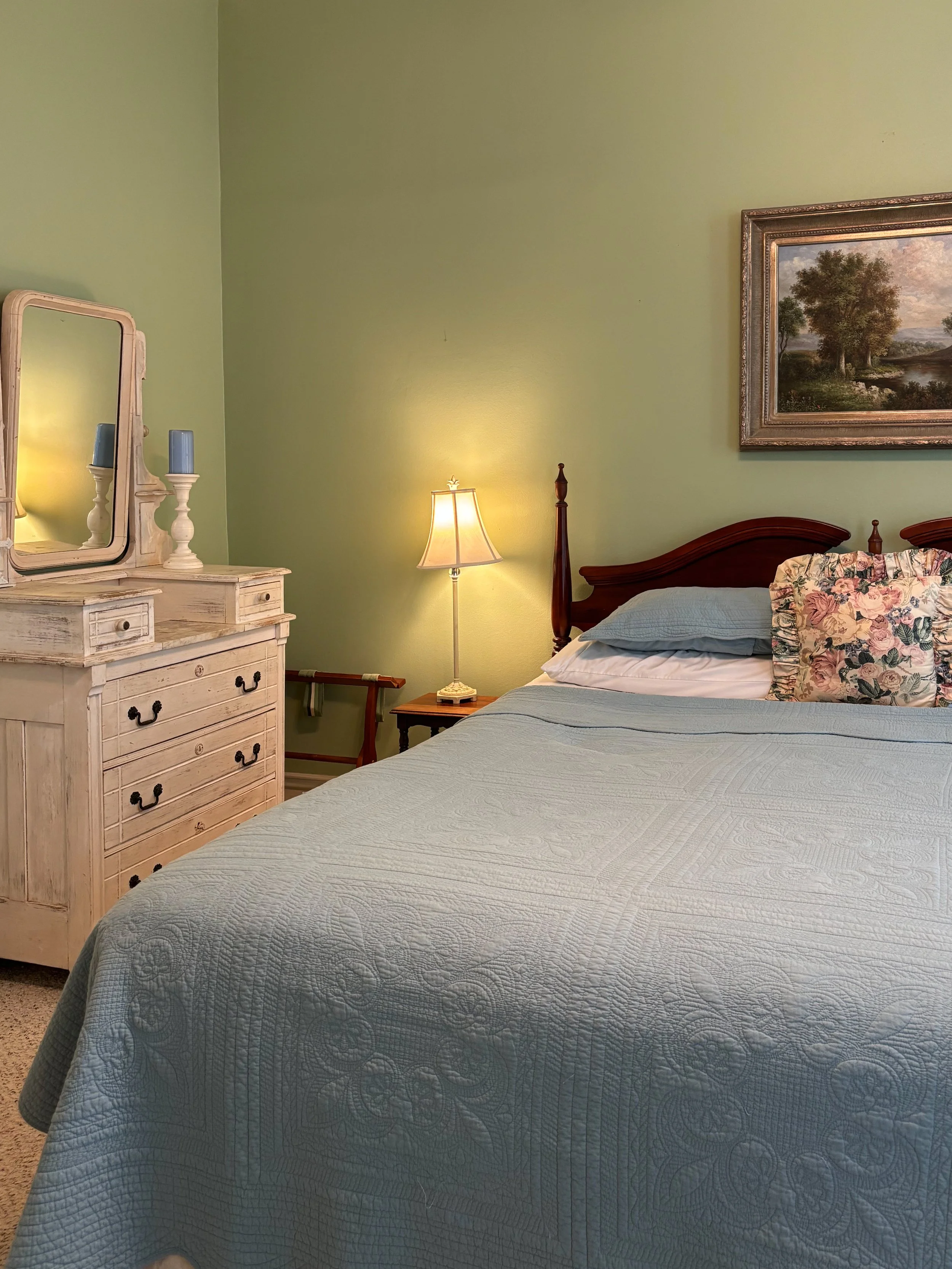 A bedroom with a bed having a quilted light gray bedspread and floral pillow, a wooden headboard, a small side table with a lit lamp, a vintage dresser with a mirror and candles, and a framed landscape painting on a green wall.
