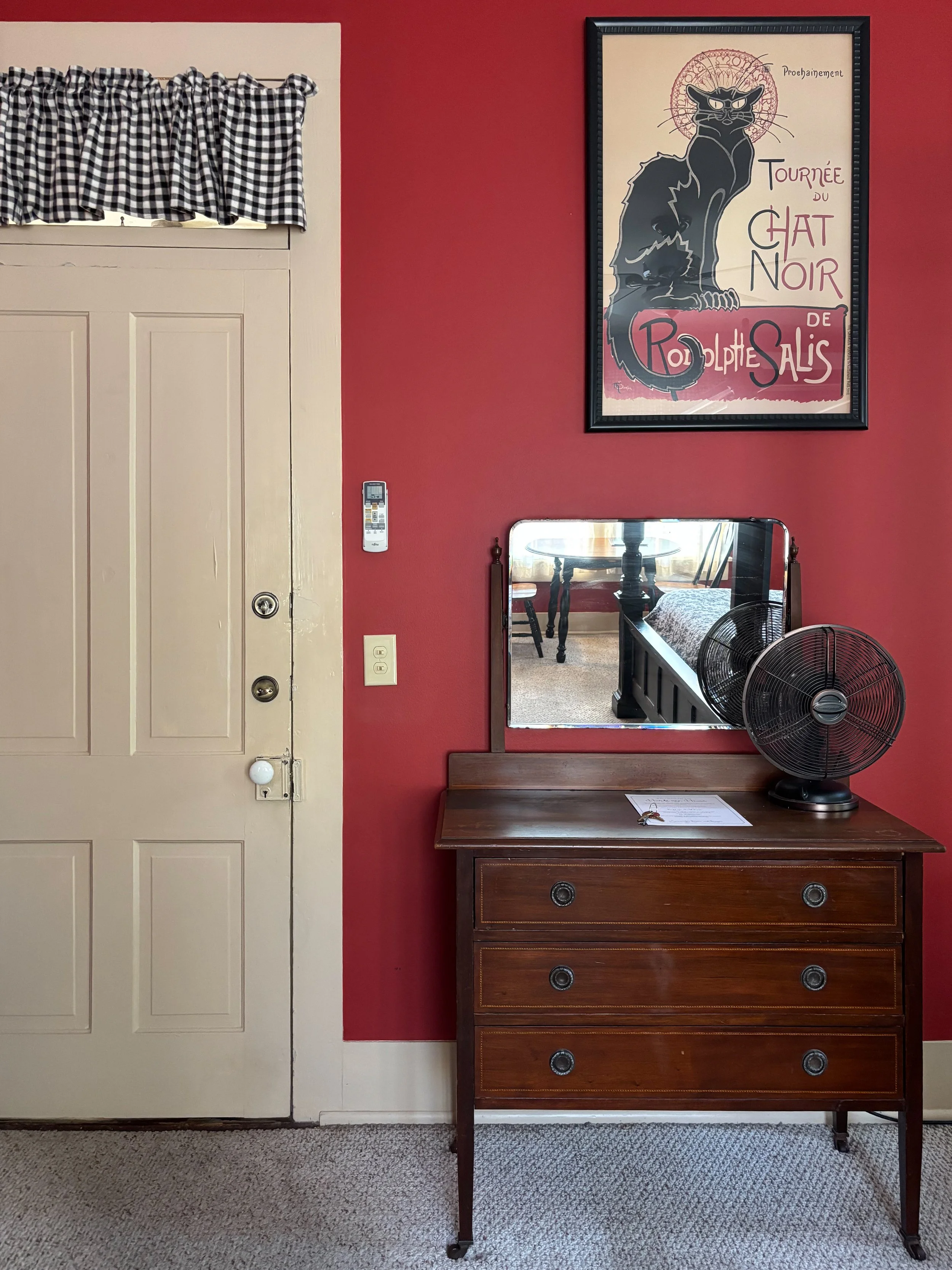 A wooden dresser with a mirror on top, a black fan, and a set of keys on it, against a red wall with framed art of a black cat and French text. A beige door with a black and white checkered valance above it is on the left side, with a remote control 
