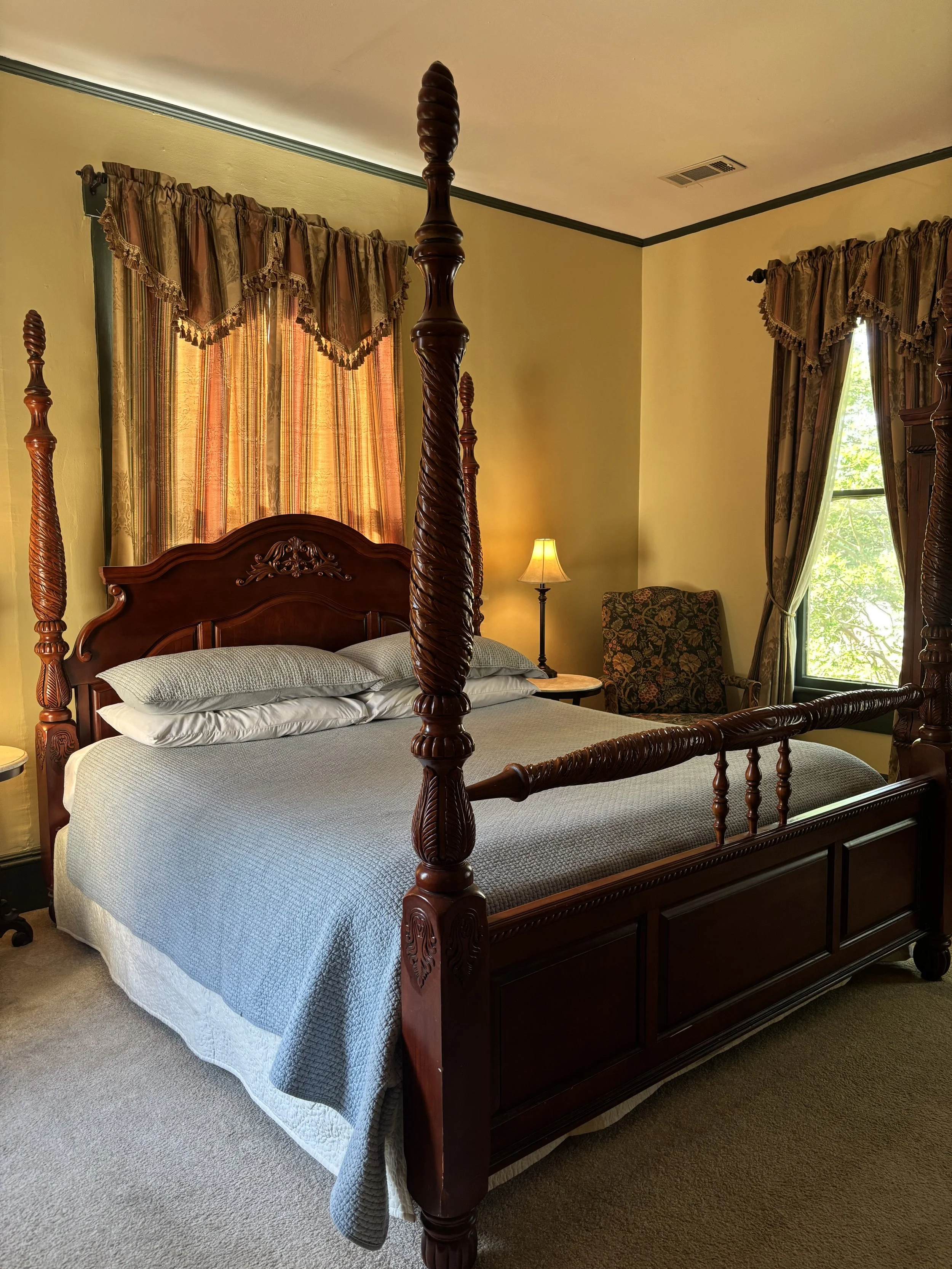A bedroom with a dark wooden four-poster bed, a blue quilt, white pillows, a side table, a table lamp, and large windows with floral and striped curtains.