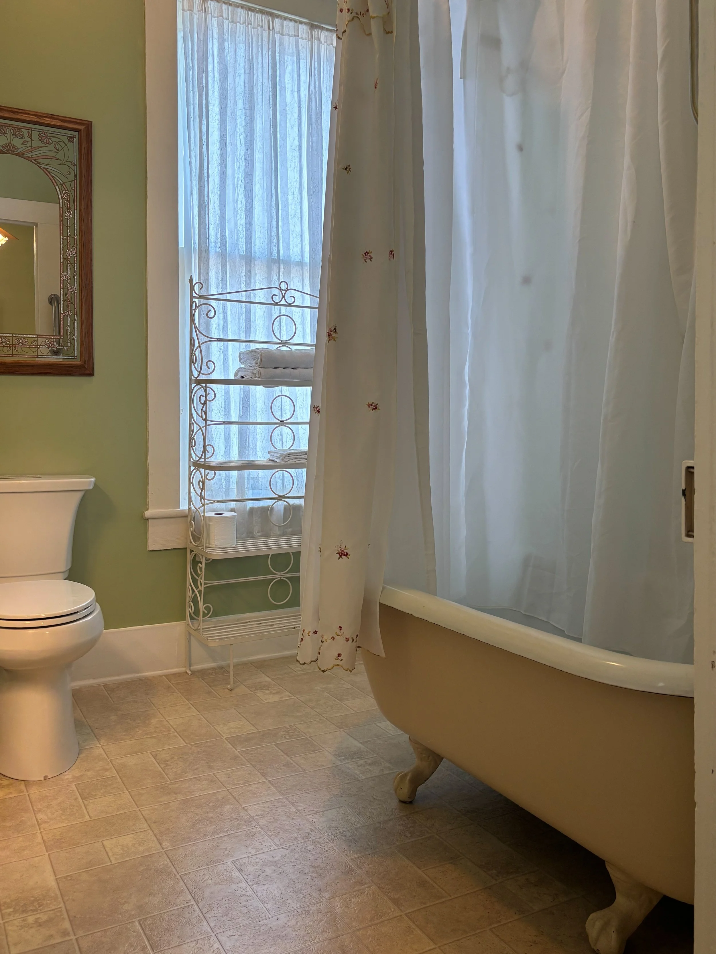 A bathroom with a vintage clawfoot bathtub, a toilet, a green wall, a mirror, a small shelf with towels, a window with curtains, and tiled flooring.