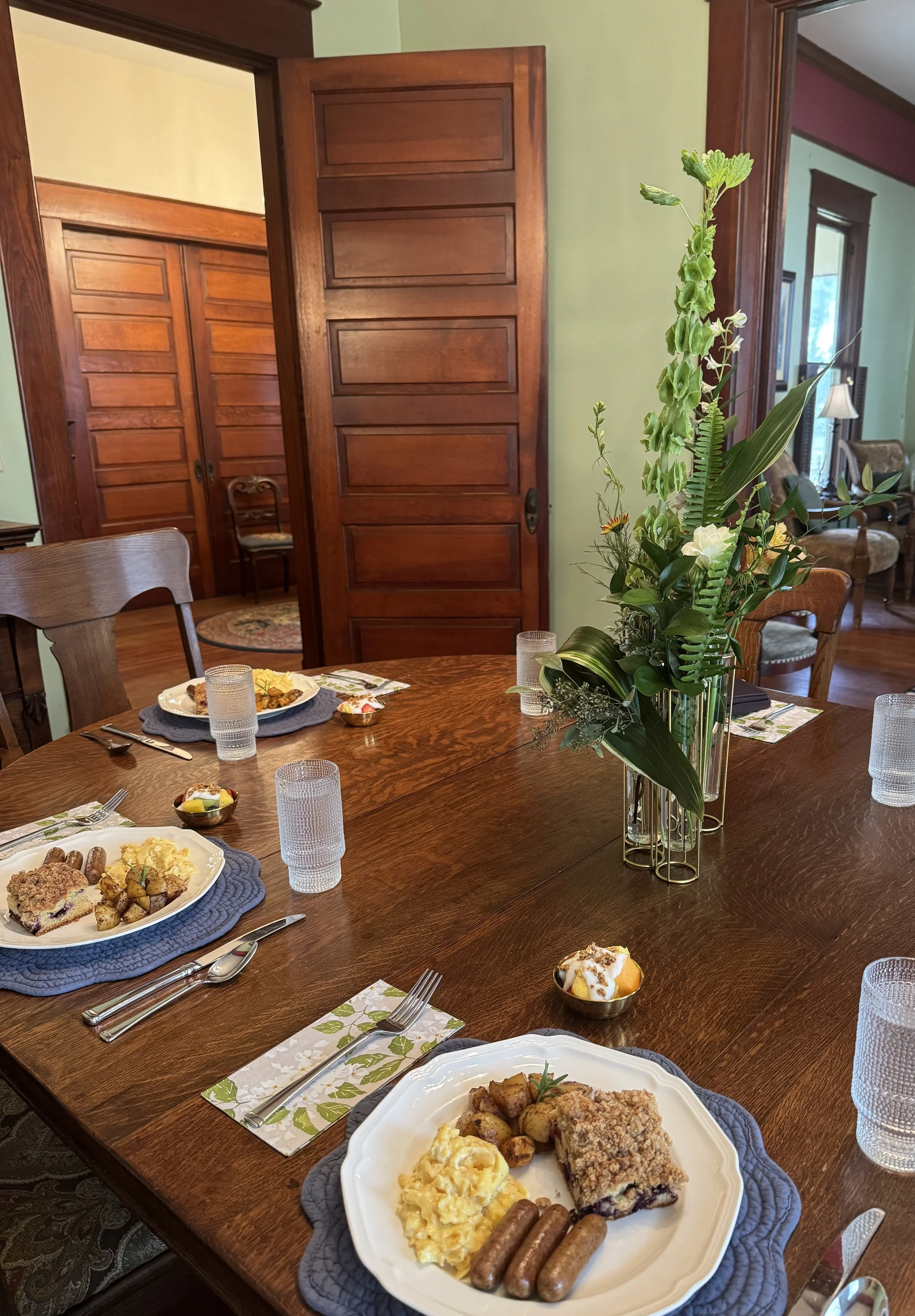 A dining room with a large wooden table set for a meal, featuring plates of food, glasses, napkins, and a centerpiece of green plants. Part of a living room with chairs and windows can be seen in the background.
