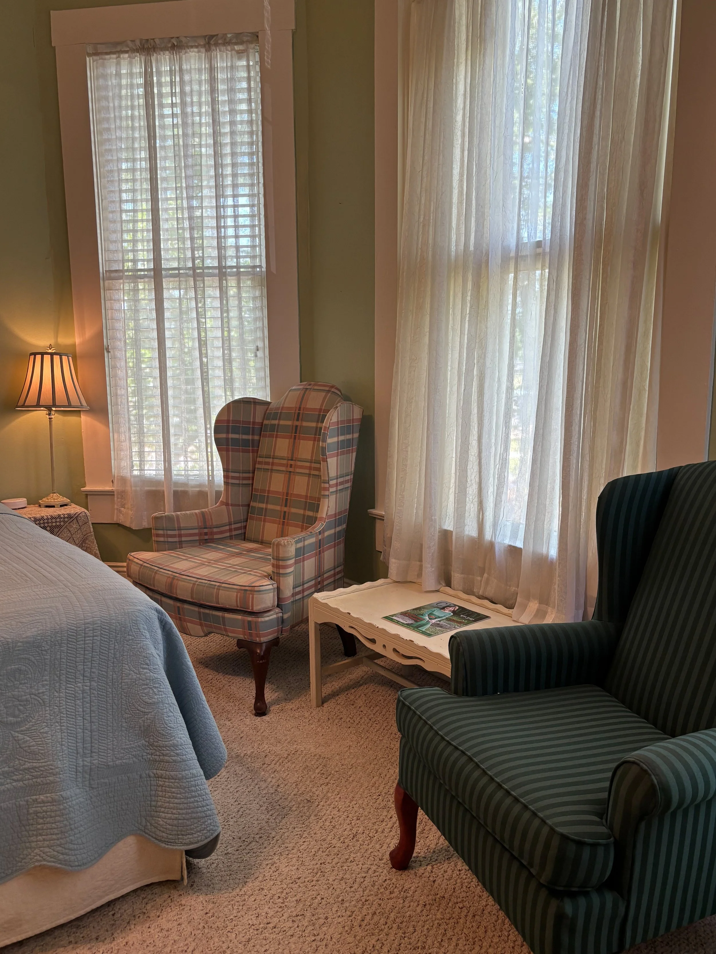 A cozy bedroom corner with two armchairs, one upholstered in plaid fabric and the other in dark green striped fabric, near windows with sheer curtains. A small white side table holds magazines, and a blue quilted bedspread is partially visible on the