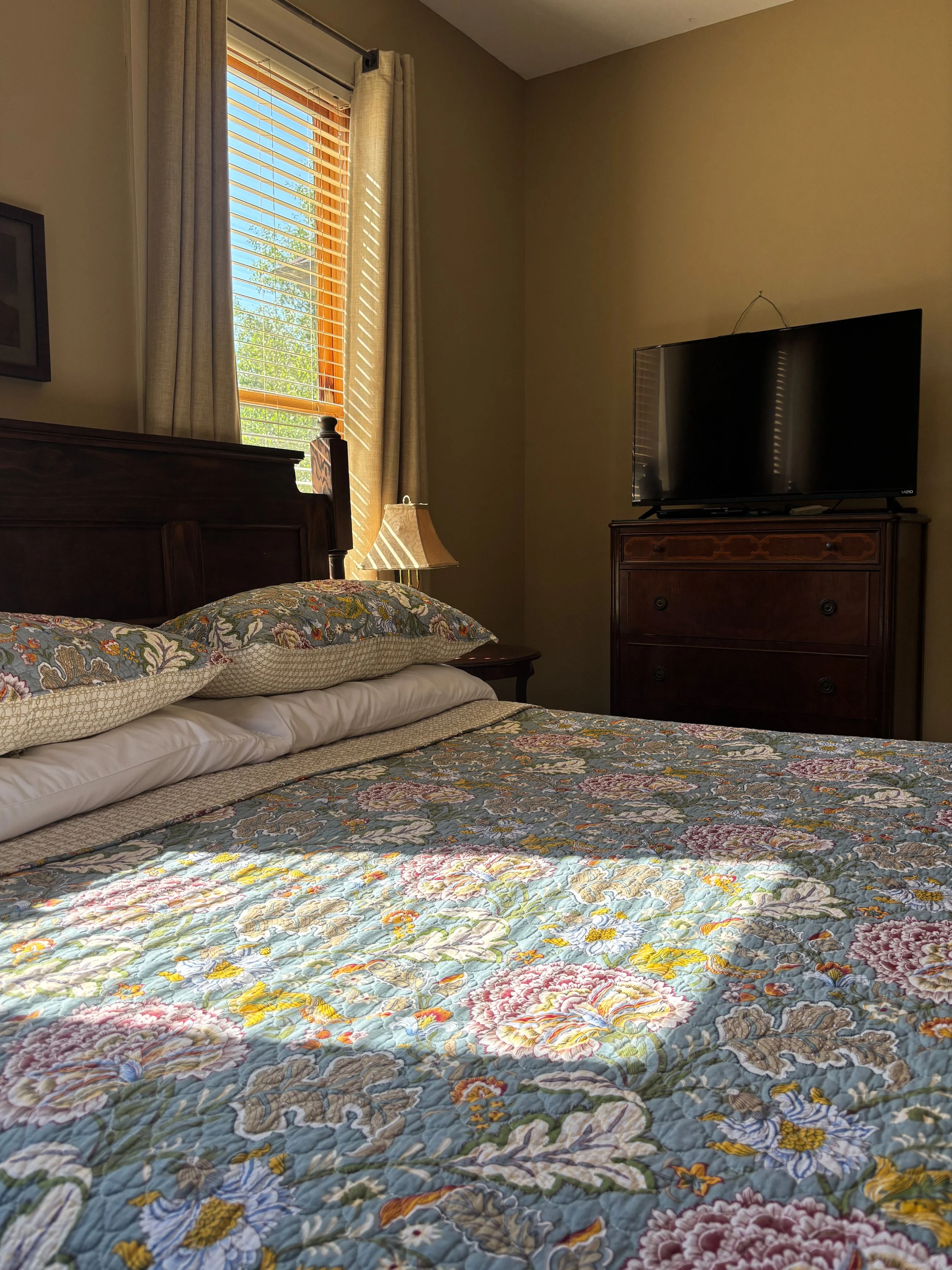 A bedroom with a bed covered by a floral quilt, pillows, a wooden headboard, a nightstand with a lamp, a window with blinds and curtains, and a television on a wooden dresser.