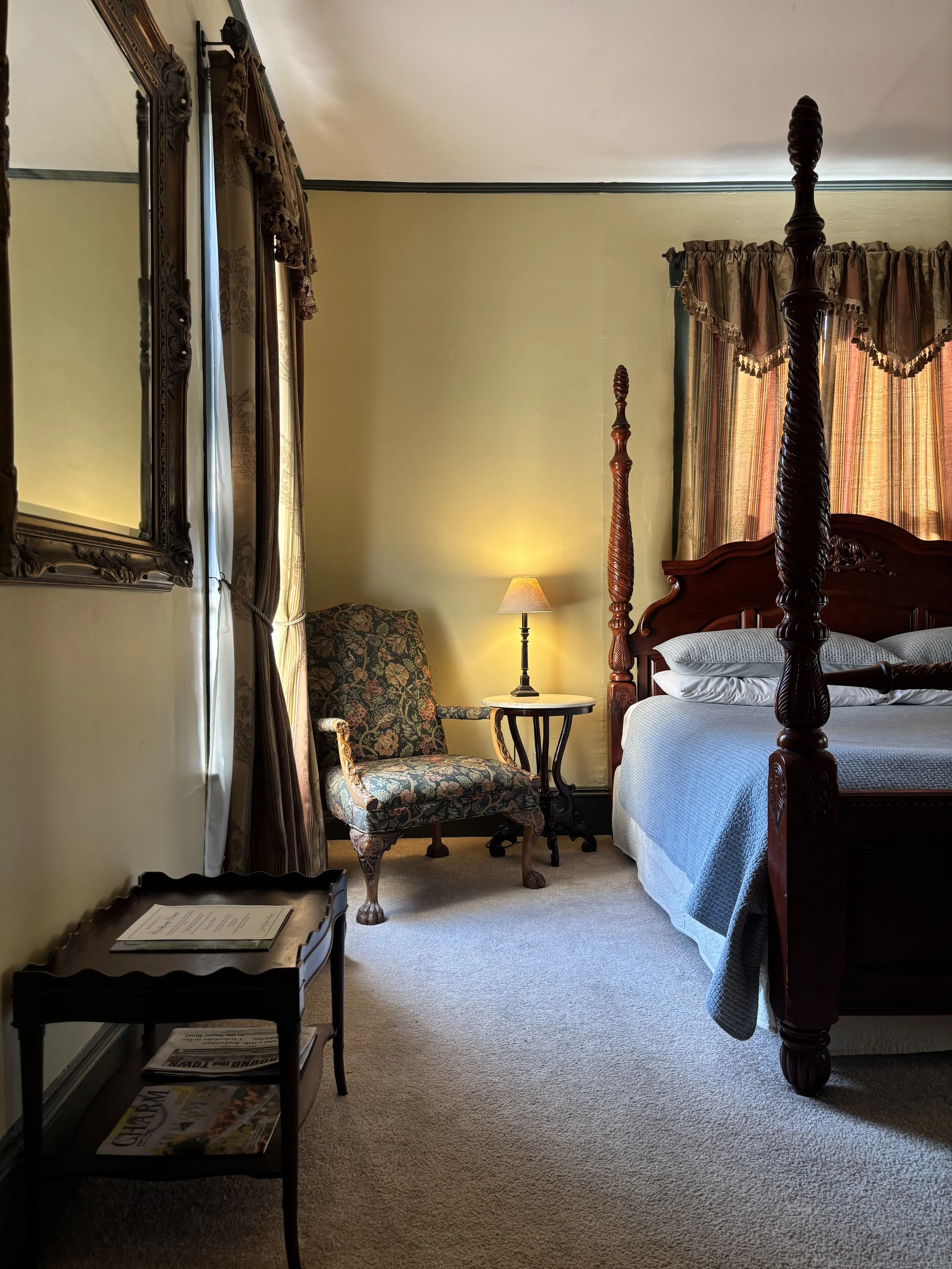 A vintage-style bedroom with a four-poster bed, floral armchair, bedside table with a lamp, wall mirror, and curtains.