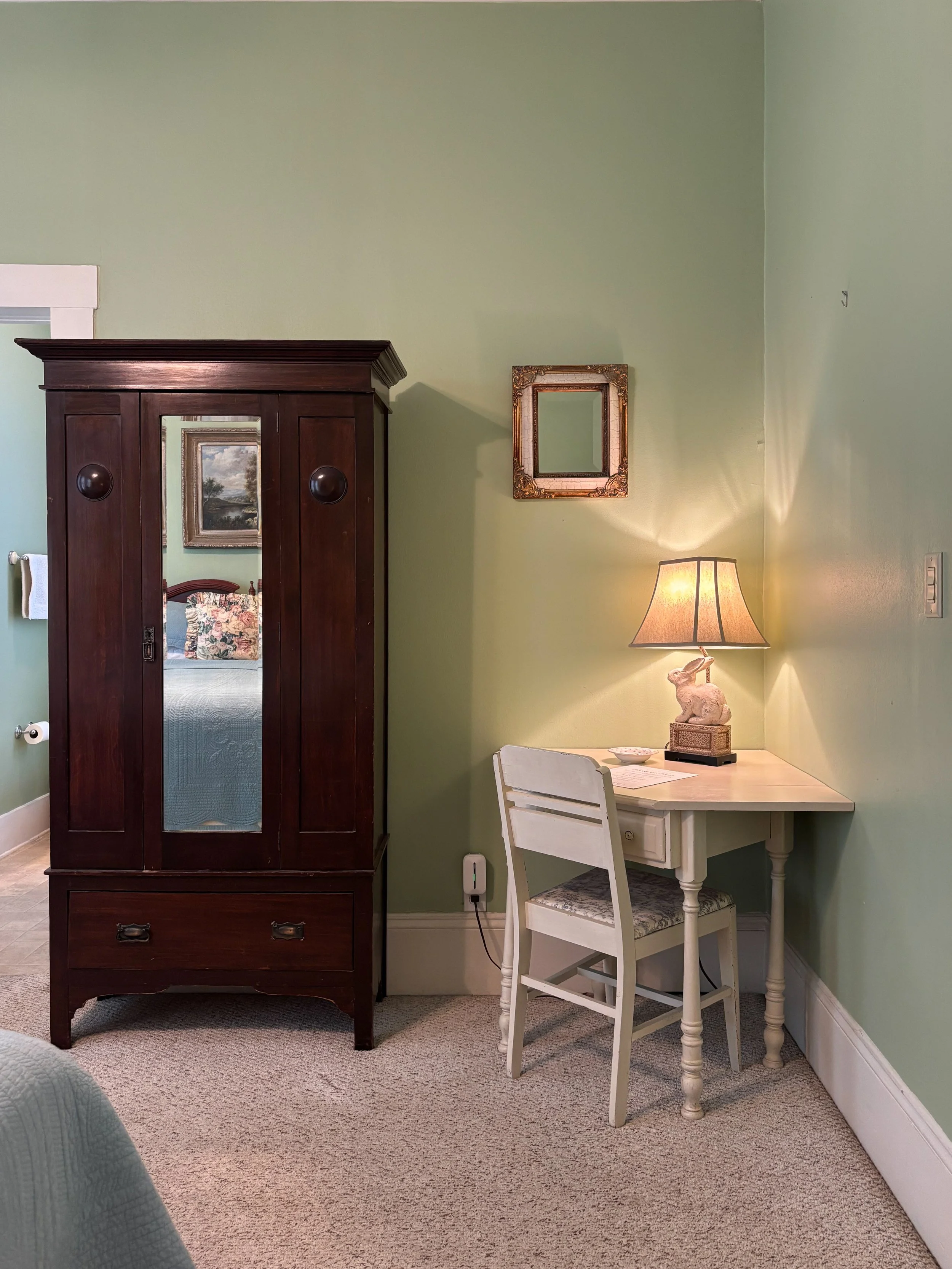 A living room corner with a dark wooden wardrobe, a small white desk with a chair, a lamp with a bunny figurine, a framed mirror, and a wall painting of a landscape, with pastel green walls and beige carpet.