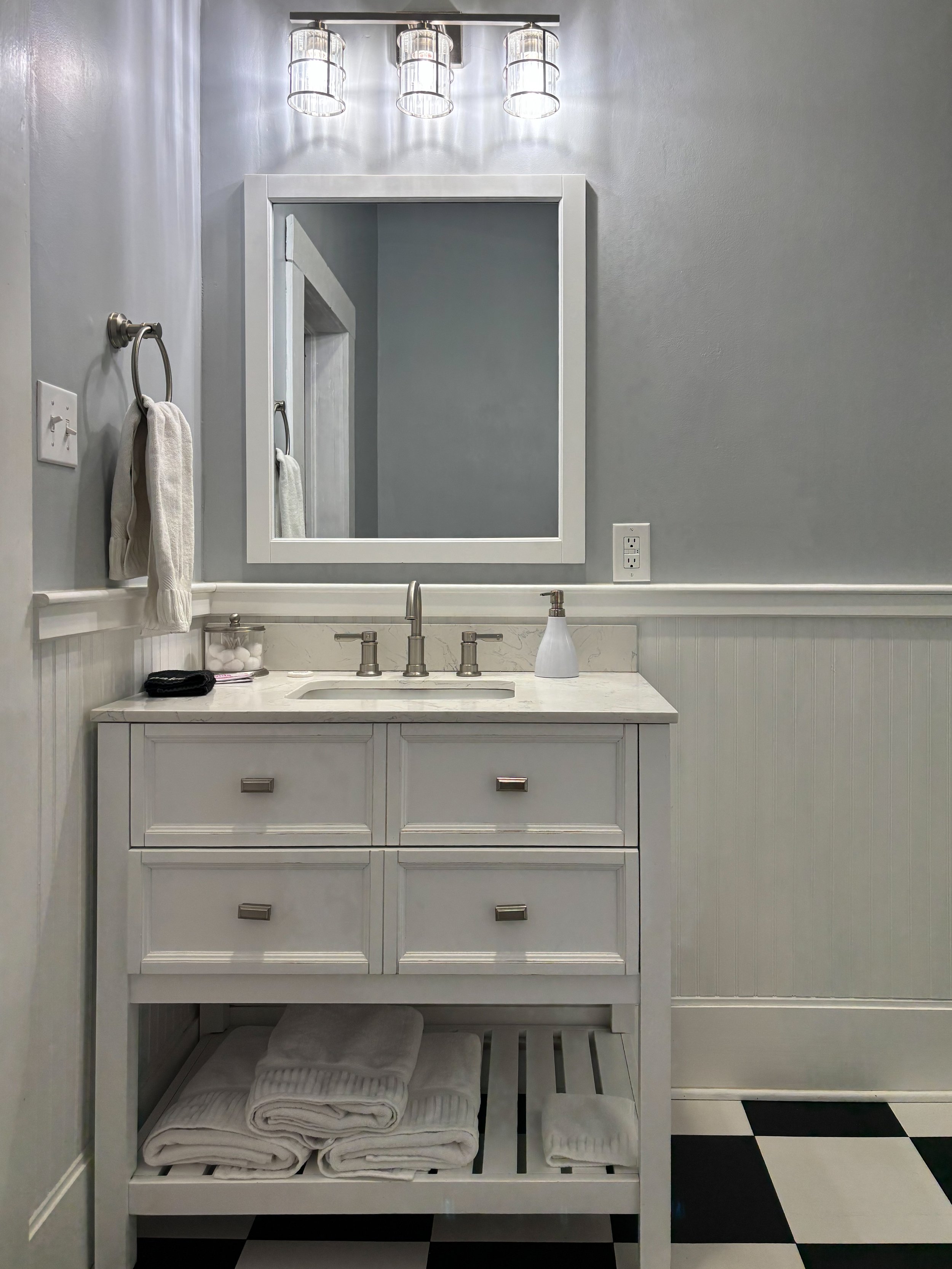 A bathroom vanity with a white wooden cabinet, three drawers, and rolled white towels underneath. A marble countertop with a faucet, soap dispenser, and a container with cotton balls. A mirror above the vanity, a soap dispenser, a towel on the ring, 
