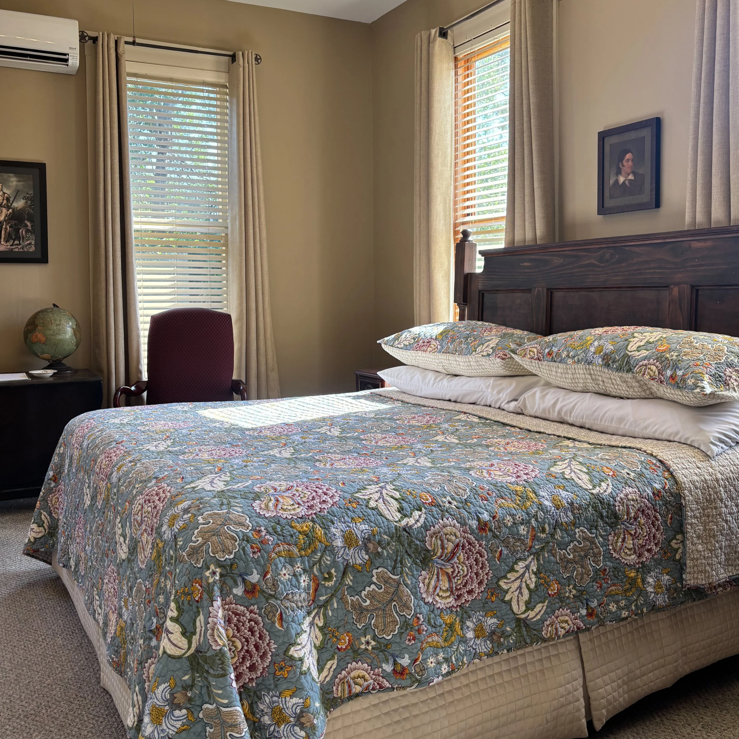 A bedroom with a bed covered in a floral quilt and pillows, wooden bed frame, beige carpet, window with blinds and curtains, framed artwork on the wall, globe on nightstand, and chair beside the bed.