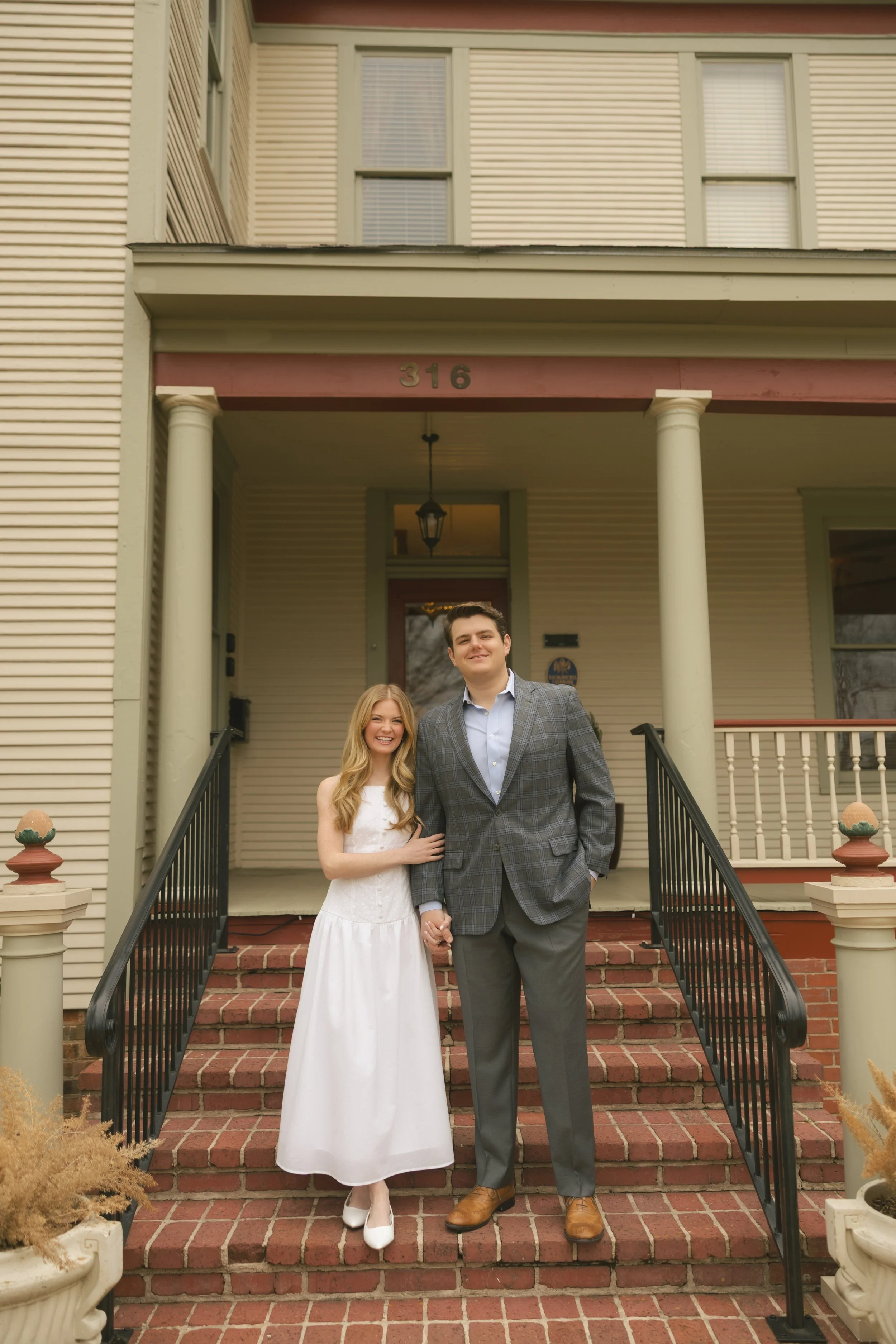 A young couple holding hands and smiling, standing on brick stairs in front of a house with cream siding, white columns, and a lantern hanging from the porch ceiling.