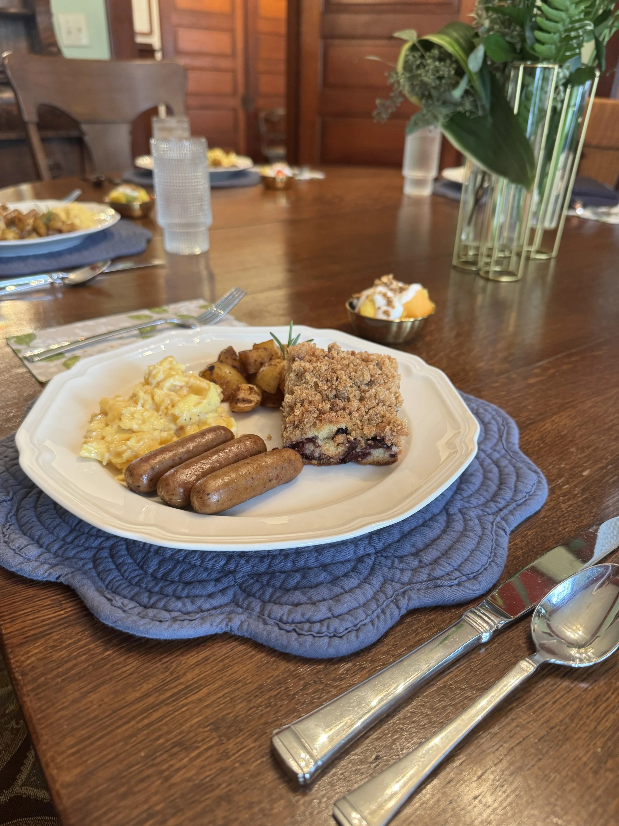 A dinner table set with a large flower arrangement in a tall metal vase, and a plate of food including scrambled eggs with cheese, sausages, roasted potatoes, and cherry pie with whipped cream and fruit topping. There are also glasses, cutlery, and p