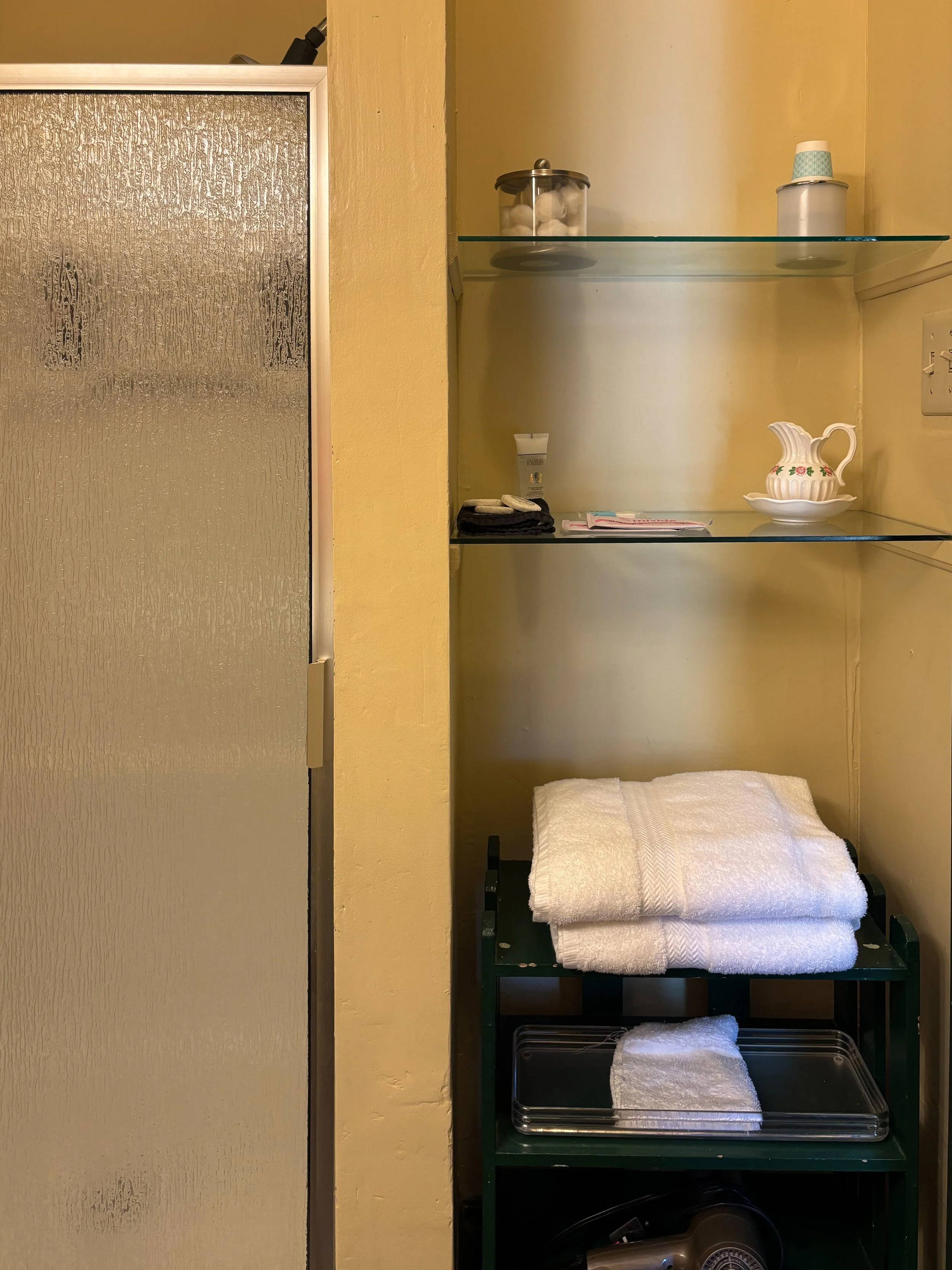 A bathroom shelf with a glass top holding cotton balls and a container of disposable cups, a porcelain creamer with a floral design, soap, folded towels, and a hairdryer.