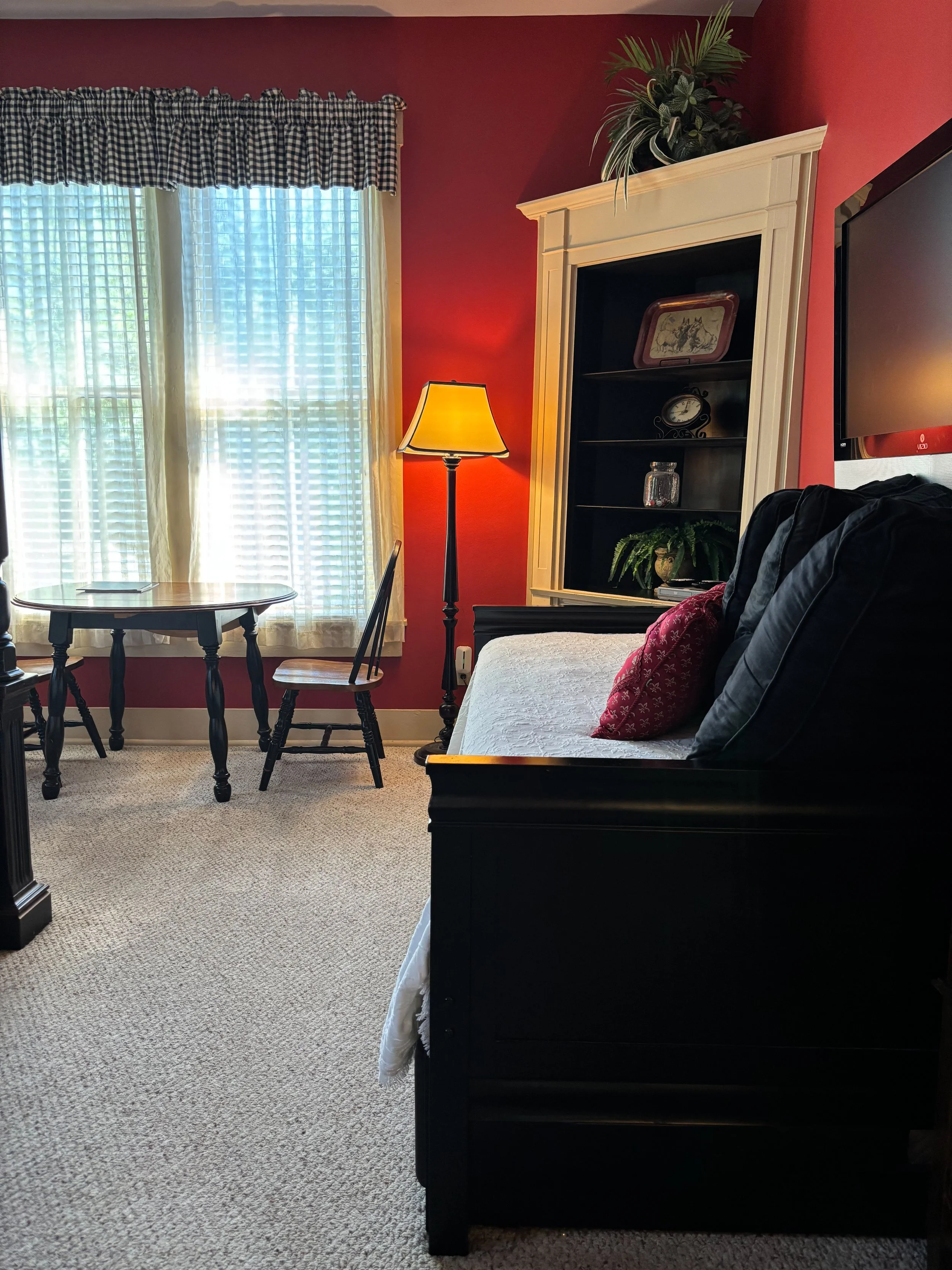 A cozy living room corner with a window featuring cream curtains and a blue checkered valance, a wooden dining table with chairs, a standing lamp, a white bookshelf with decorative items and plants, a wall-mounted TV, and a bed with black pillows and