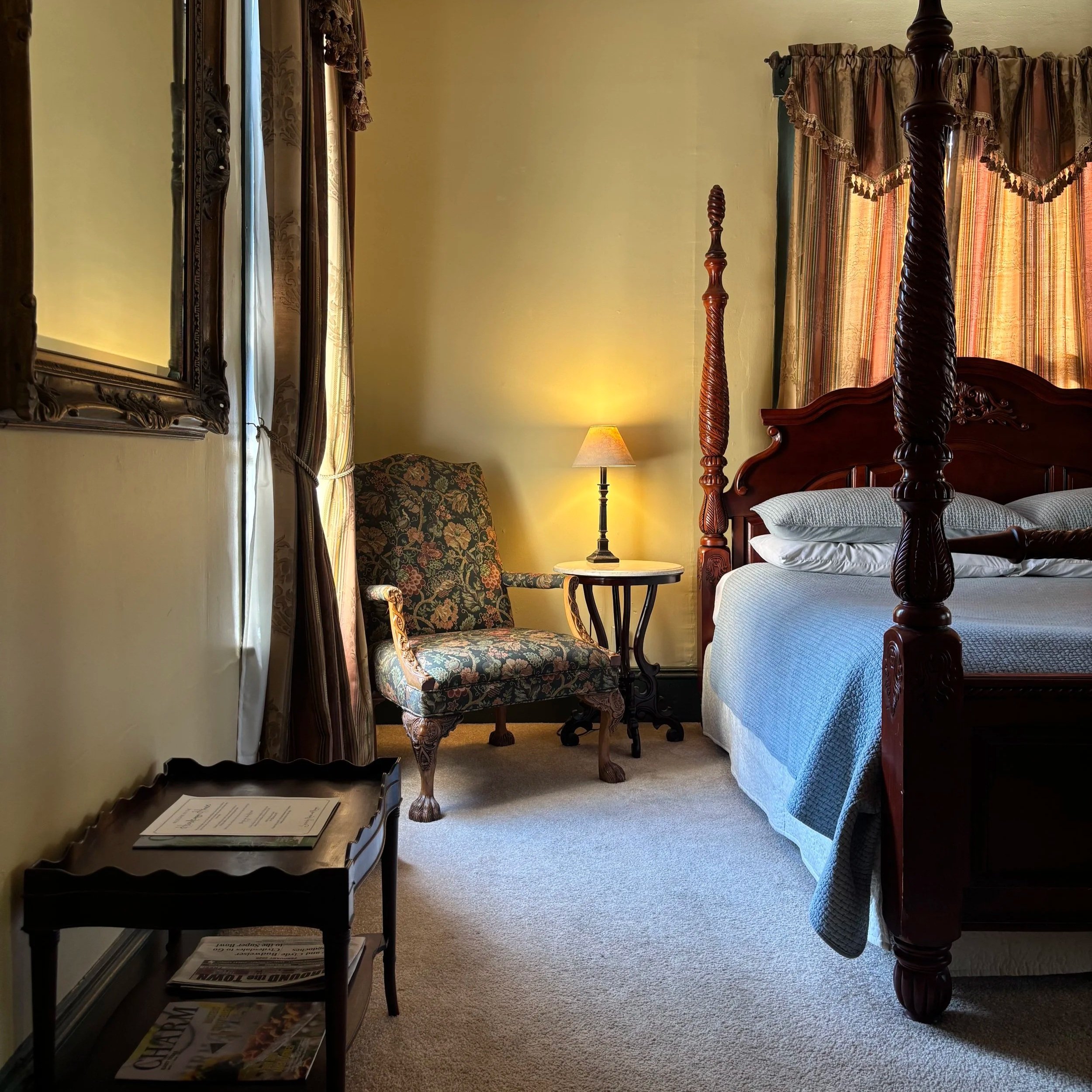 A cozy hotel room with a wooden four-poster bed, floral patterned armchair, lamp, mirror, and window with floral curtains.