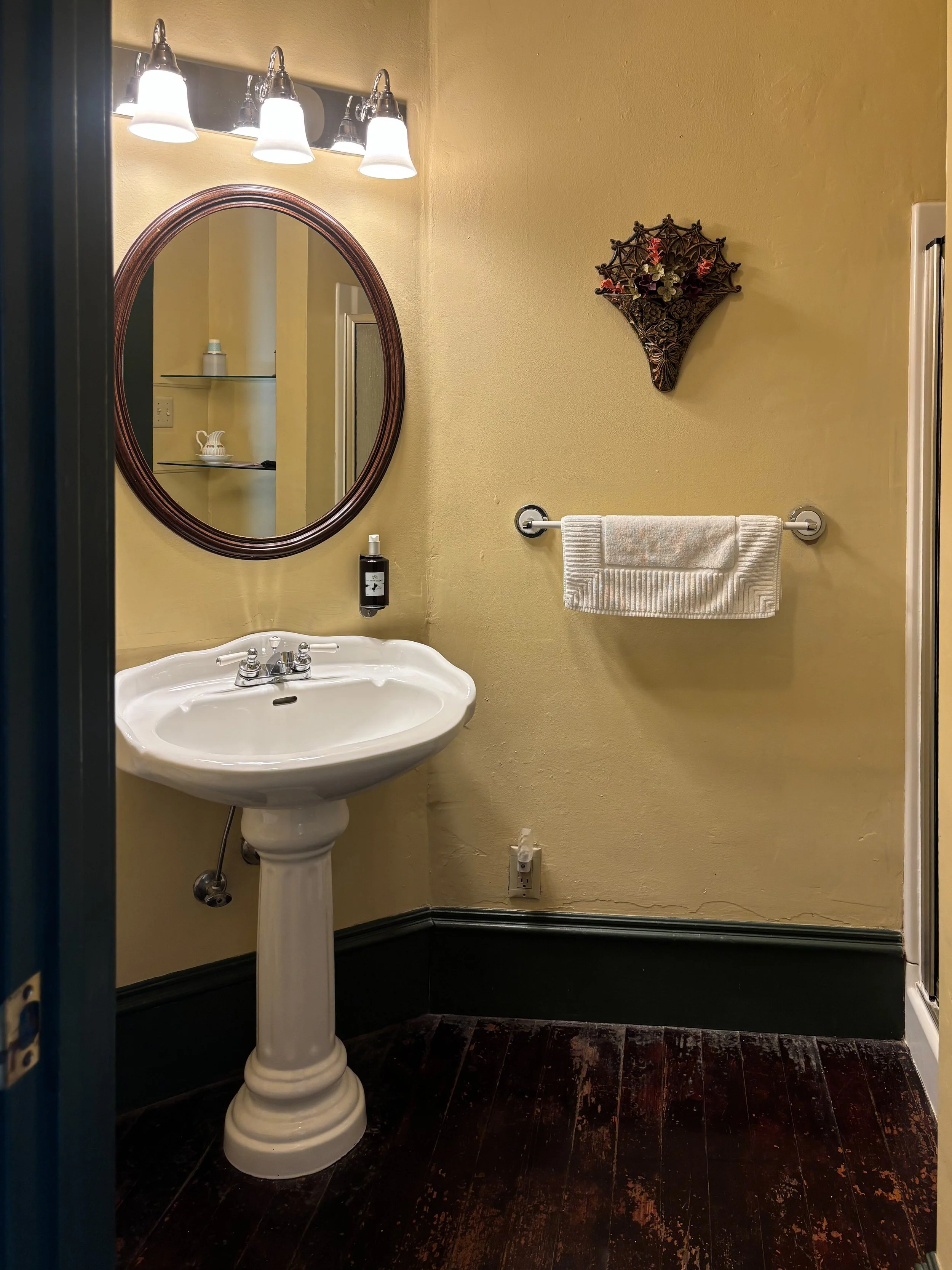 A small bathroom with a white pedestal sink, a mirror with a wooden frame, a wall-mounted light fixture with four lights, a towel rack with a white towel, and decorative wall art. The walls are painted yellow, and the floor is dark wood.