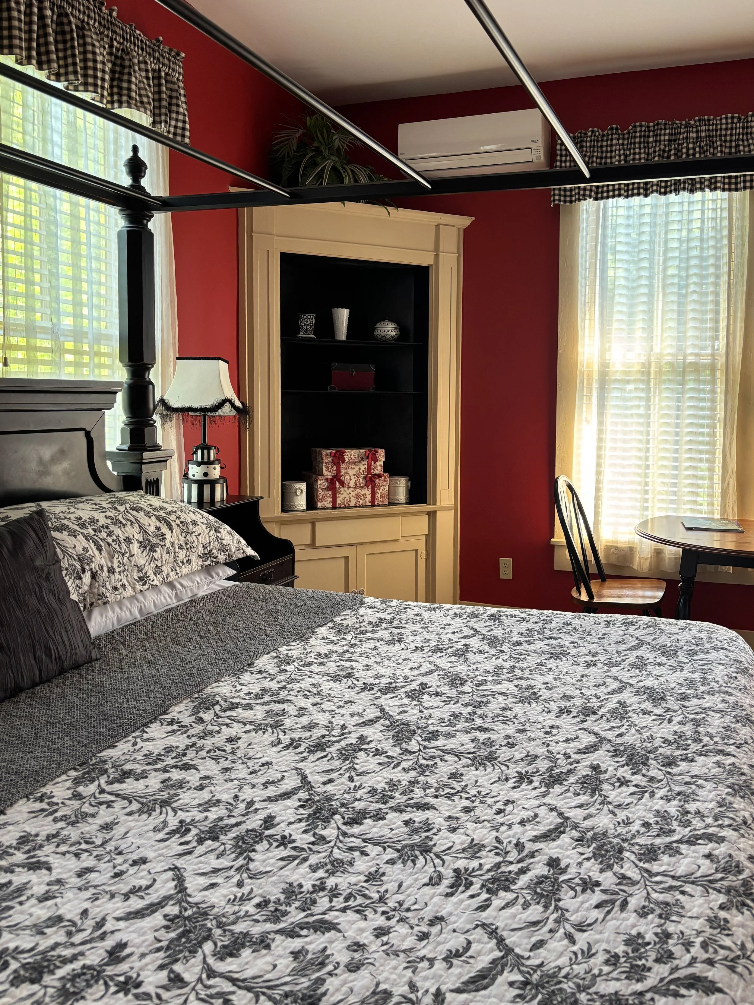 A bedroom with a bed covered in a black and white floral quilt, black records, and pillows. A nightstand with a black and white lamp is beside the bed. In the background, there's a cream-colored bookshelf with decorative items and wrapped gifts, a wi