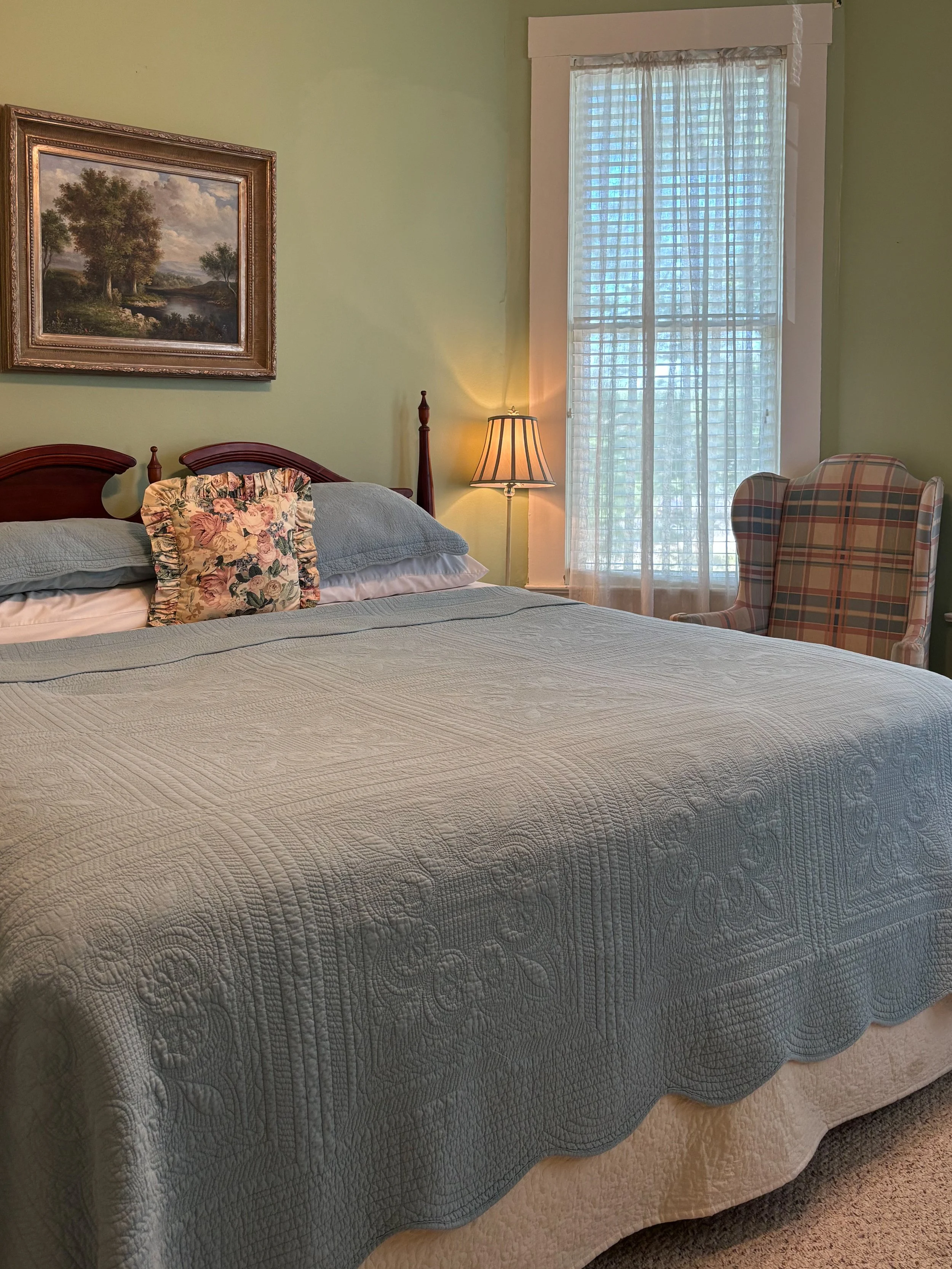 A neatly made bed with a white quilt cover, a floral decorative pillow, and light blue pillows, in a bedroom with a green wall, a framed landscape painting, a table lamp, and a window with white curtains and blinds.